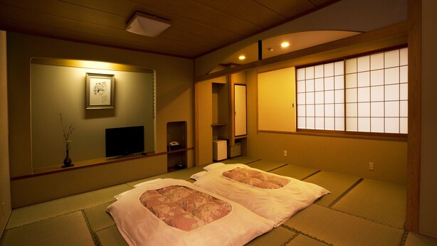 Una habitación japonesa tradicional con esteras de tatami, dos futones de tatami con cojines rosas, un televisor, un espejo y un biombó shoji.