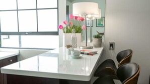 A modern kitchen with a white countertop, a glass-top island, and black chairs. There is a potted plant with pink flowers and a lamp on the island. - FlowSuites Polanco (Mexico City)