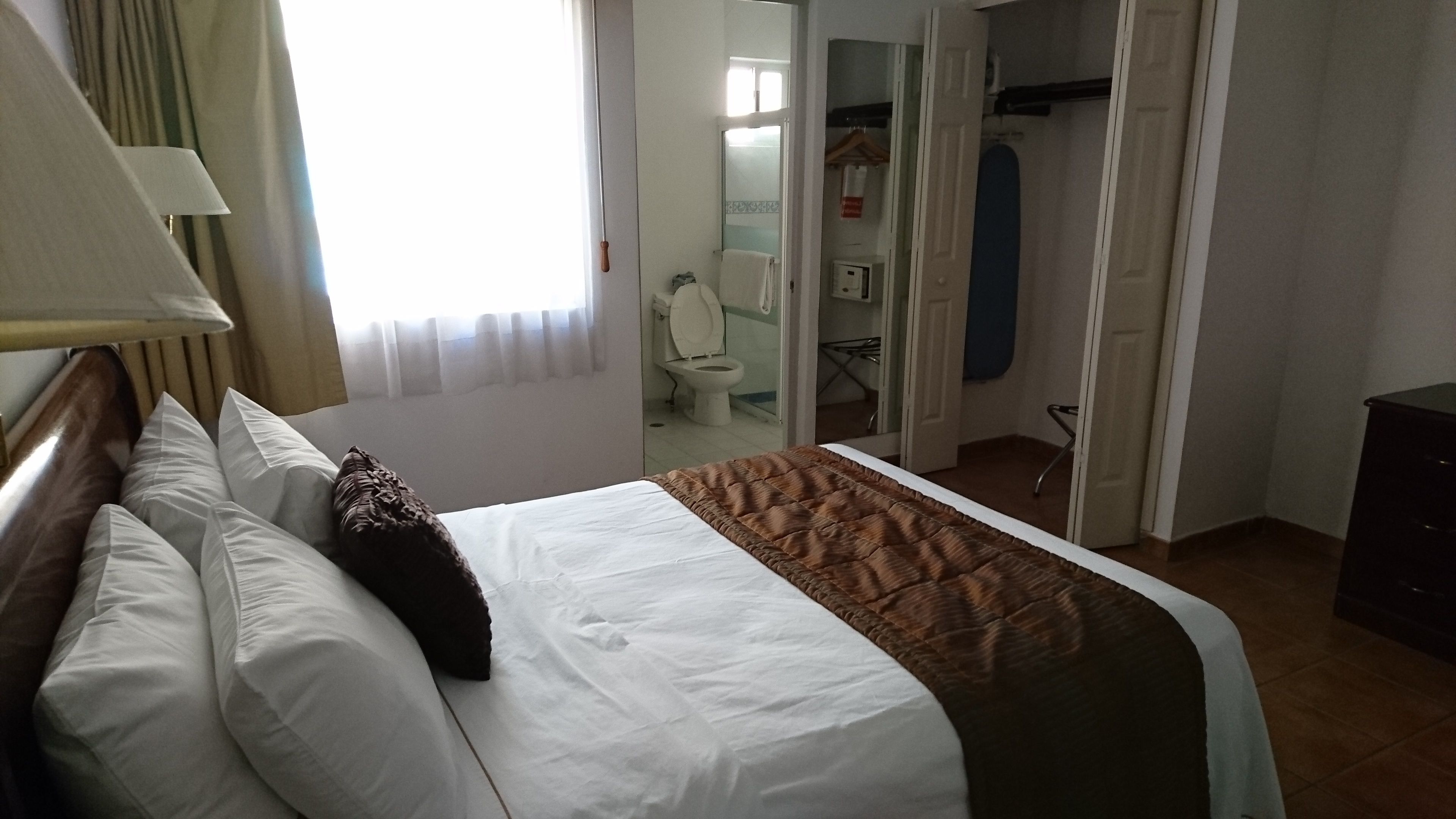 Habitación de hotel con cama, mesita de noche, lámpara, ventana con cortinas, baño visible a través de una puerta abierta y perchero.