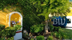 A yellow building with a white archway entrance, a garden with various plants, and a blue vehicle with the letters "BW" on it. - Best Western Saltillo (Saltillo)