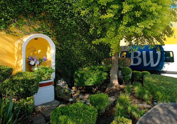 Un edificio amarillo con un arco de entrada blanco, un jardín con diversas plantas y un vehículo azul con las letras "BW".