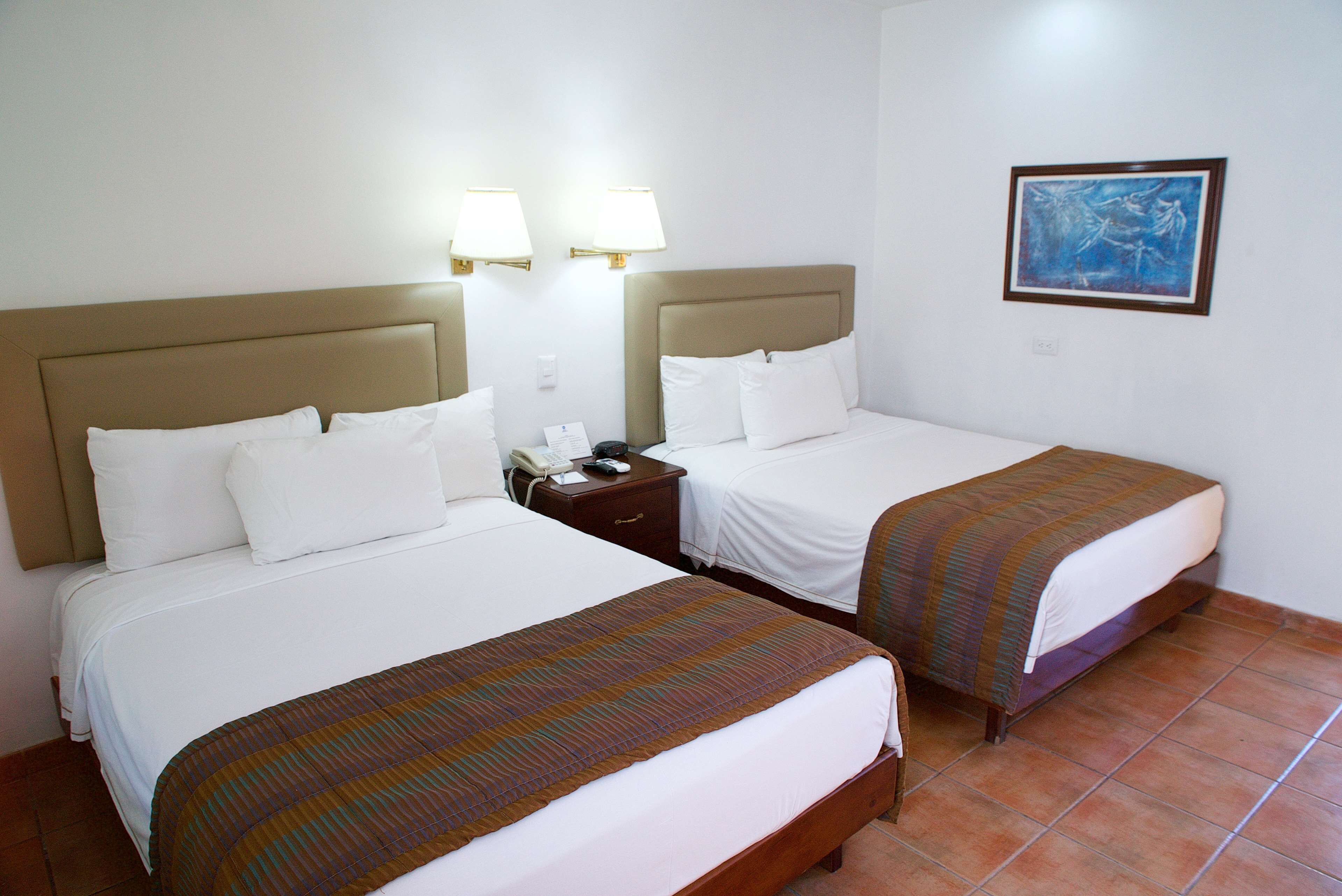 Habitación de hotel con dos camas, cada una con cabecera, mesita de noche con teléfono y un cuadro en la pared.