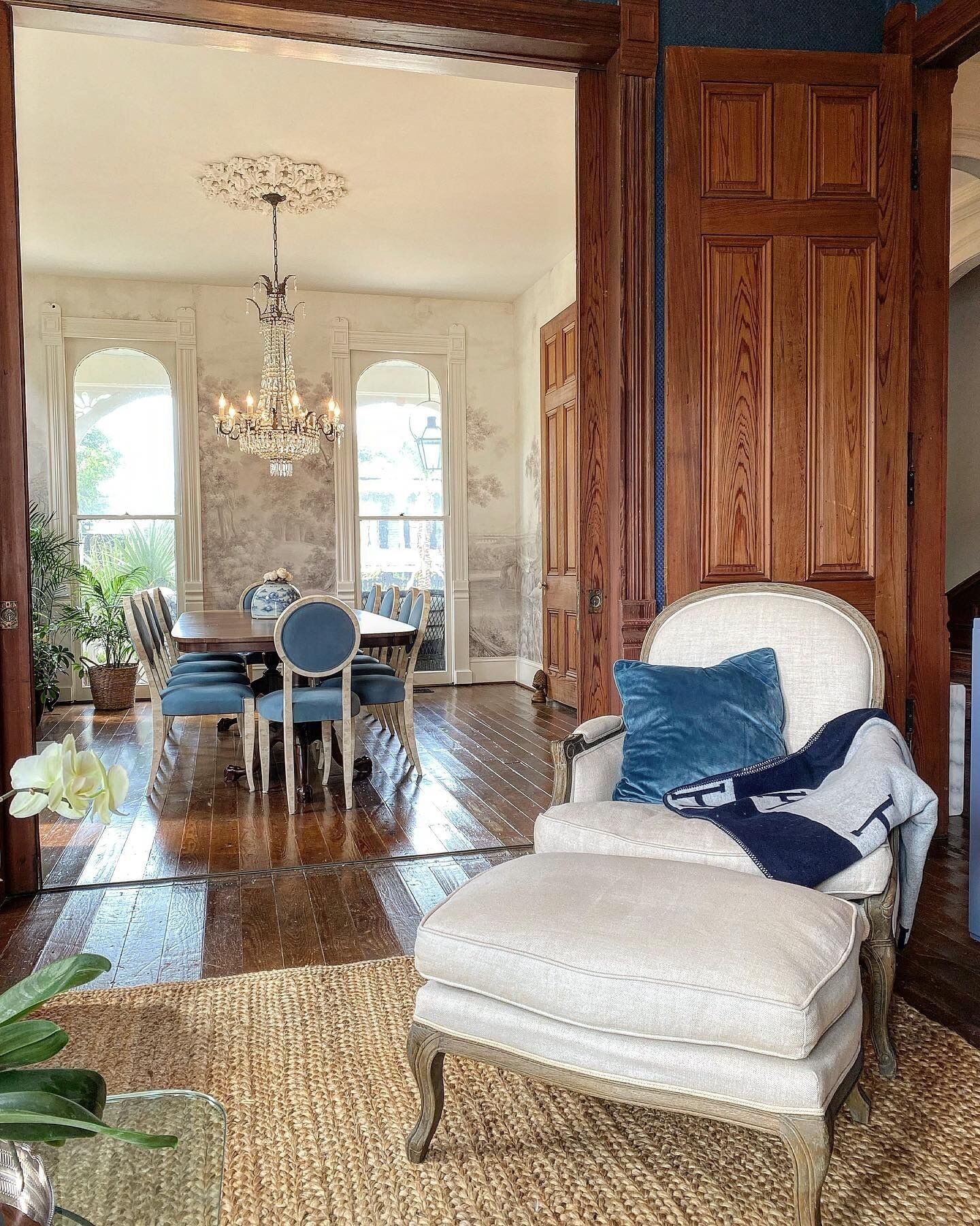 A spacious living room with a large white armchair, a dining area with blue chairs, and a chandelier.