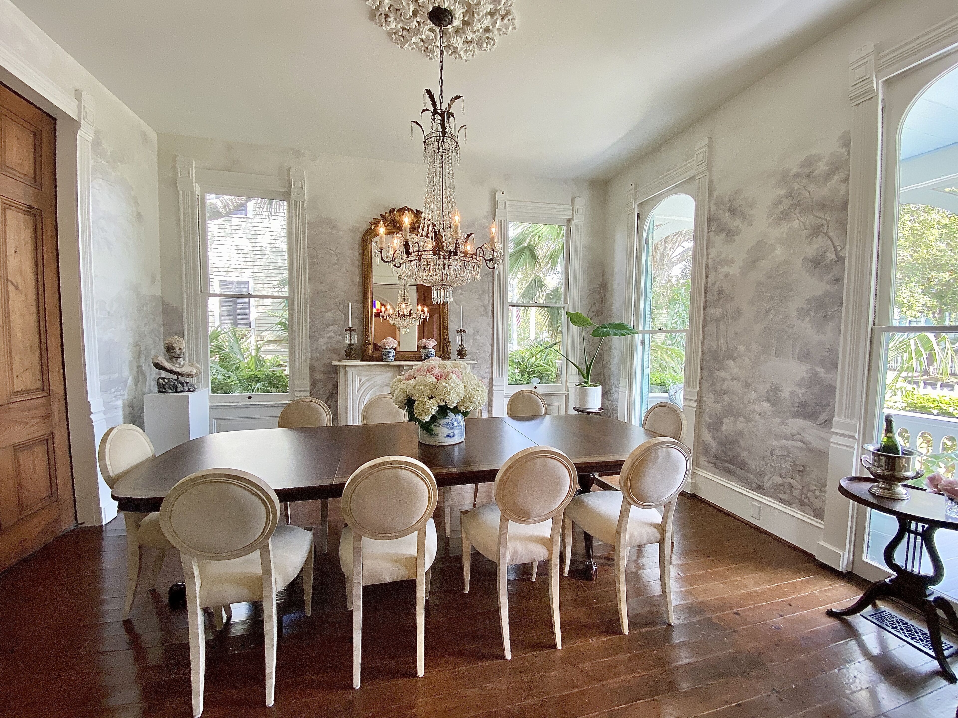 A dining room with a large wooden table, six white upholstered chairs, a chandelier, and a view of greenery outside.