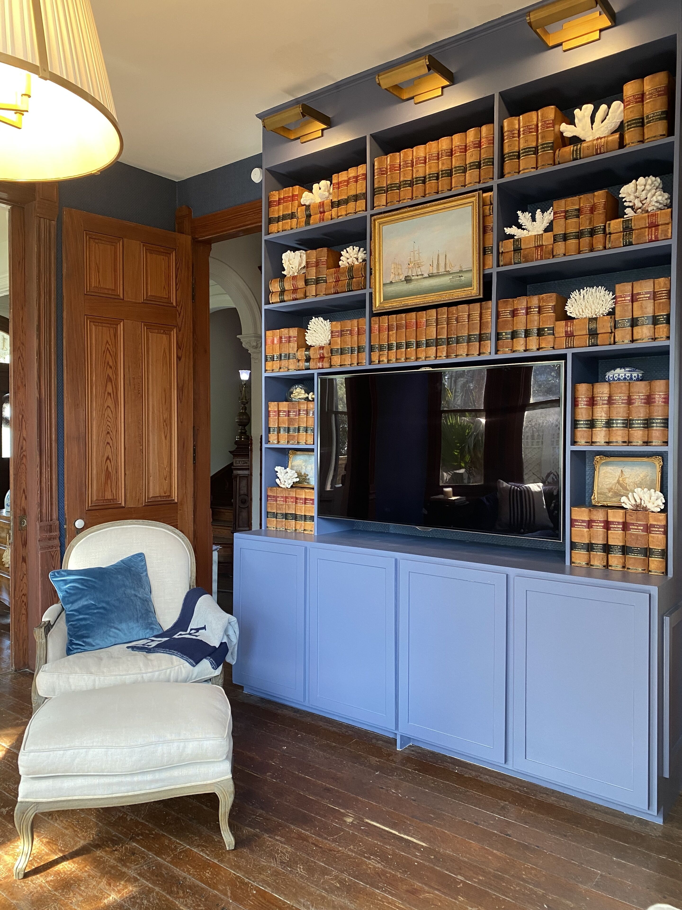 A room with a wooden door, a white armchair with a blue pillow, and a bookshelf filled with books and decorative items.