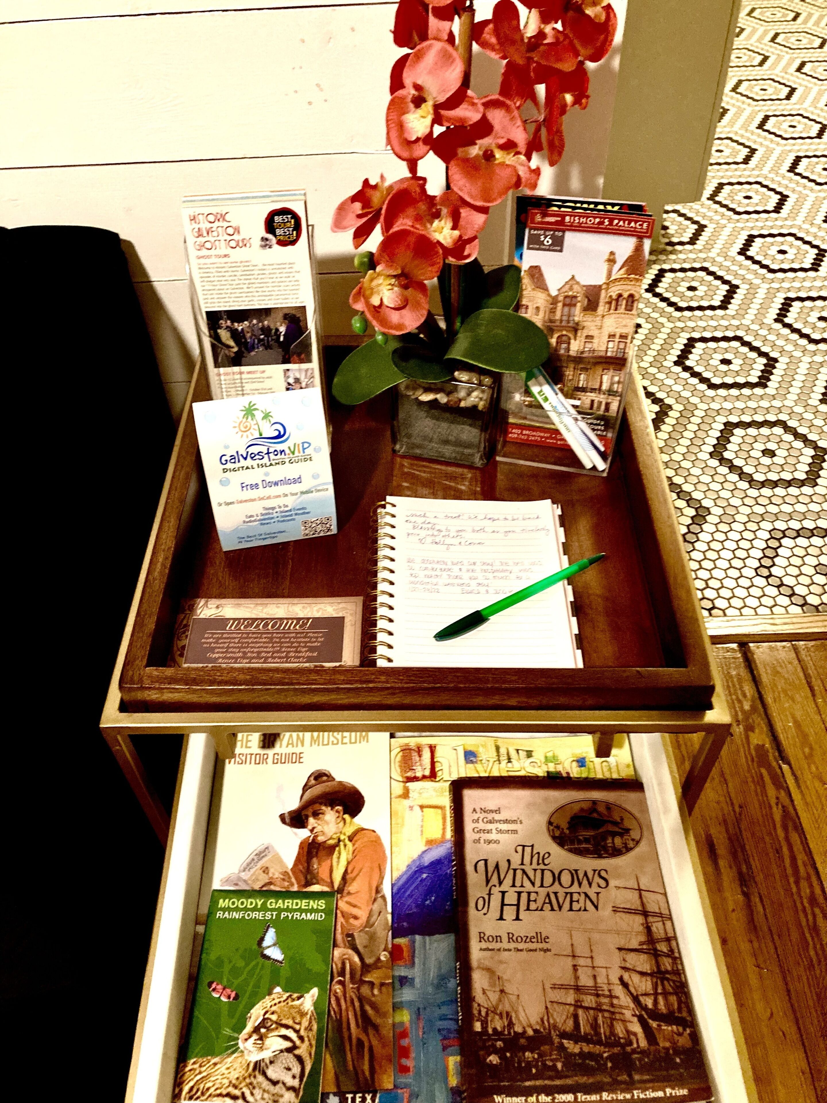 A wooden table with a variety of items including a digital island guide, a visitor guide, a book titled 'The Windows of Heaven,' and a notebook with a pen.