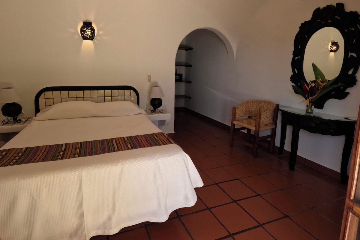 A bedroom with a bed, bedside lamps, a chair, a mirror, and a shelf with decorative items.