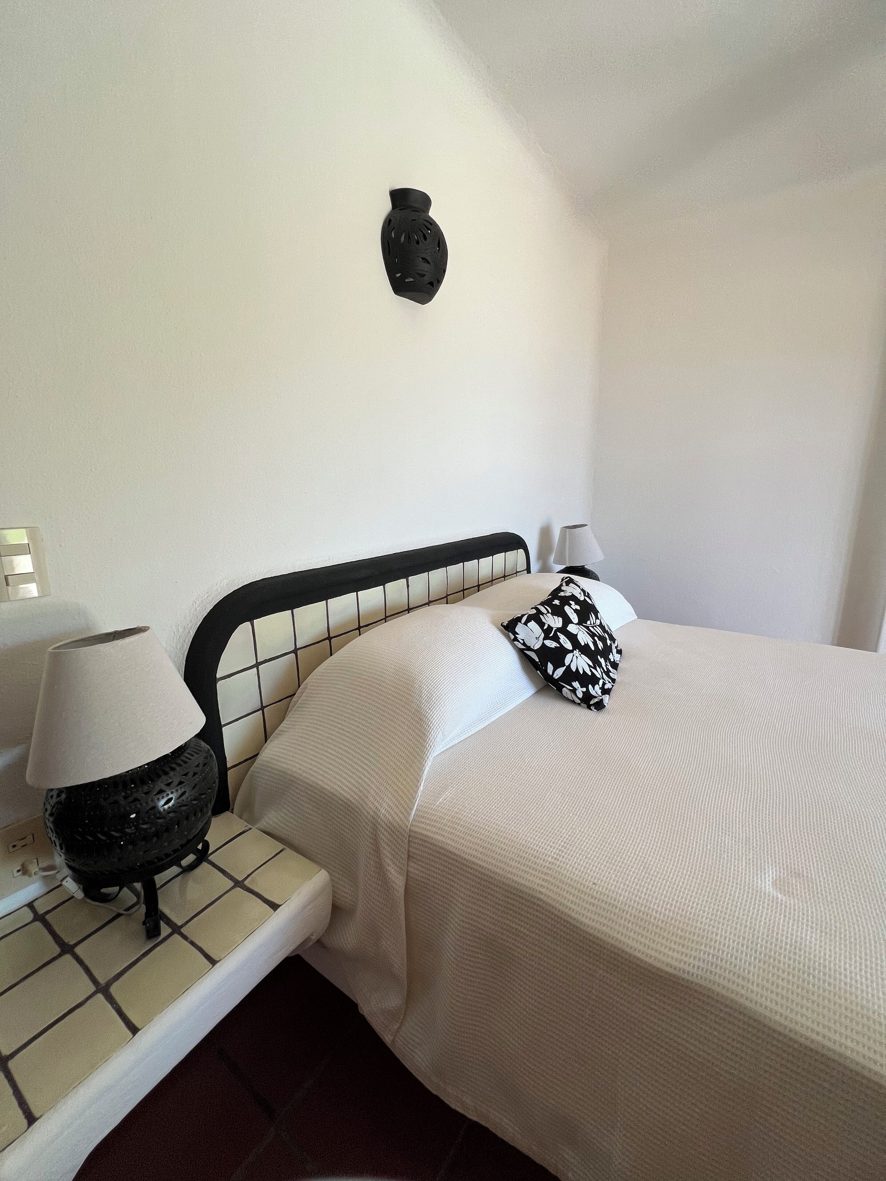 A bed with a black metal headboard, a white pillow, a black and white patterned pillow, and a black lamp on a bedside table.