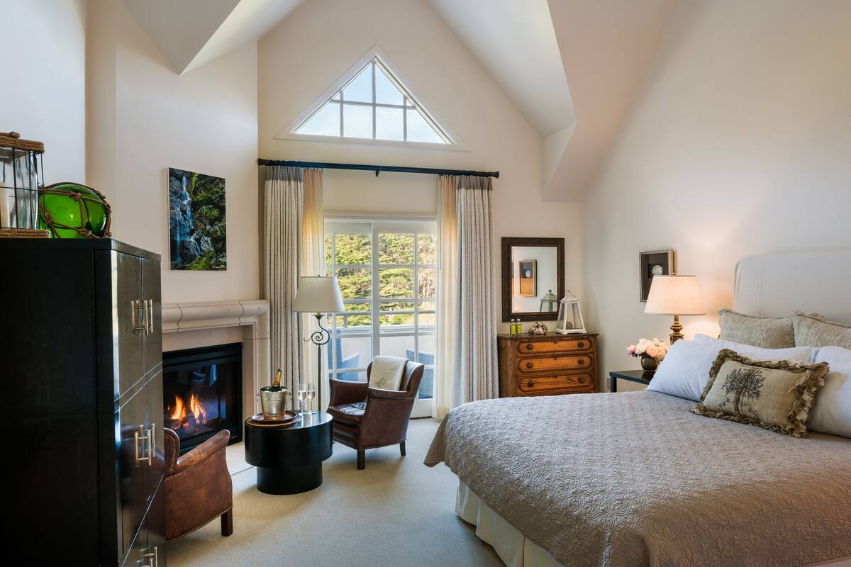 A spacious bedroom with a high ceiling, a fireplace, a large bed, a nightstand, a dresser, and a view of the outdoors through a window.