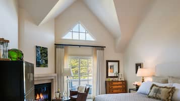 A spacious bedroom with a high ceiling, a fireplace, a large bed, a nightstand, a dresser, and a view of the outdoors through a window.