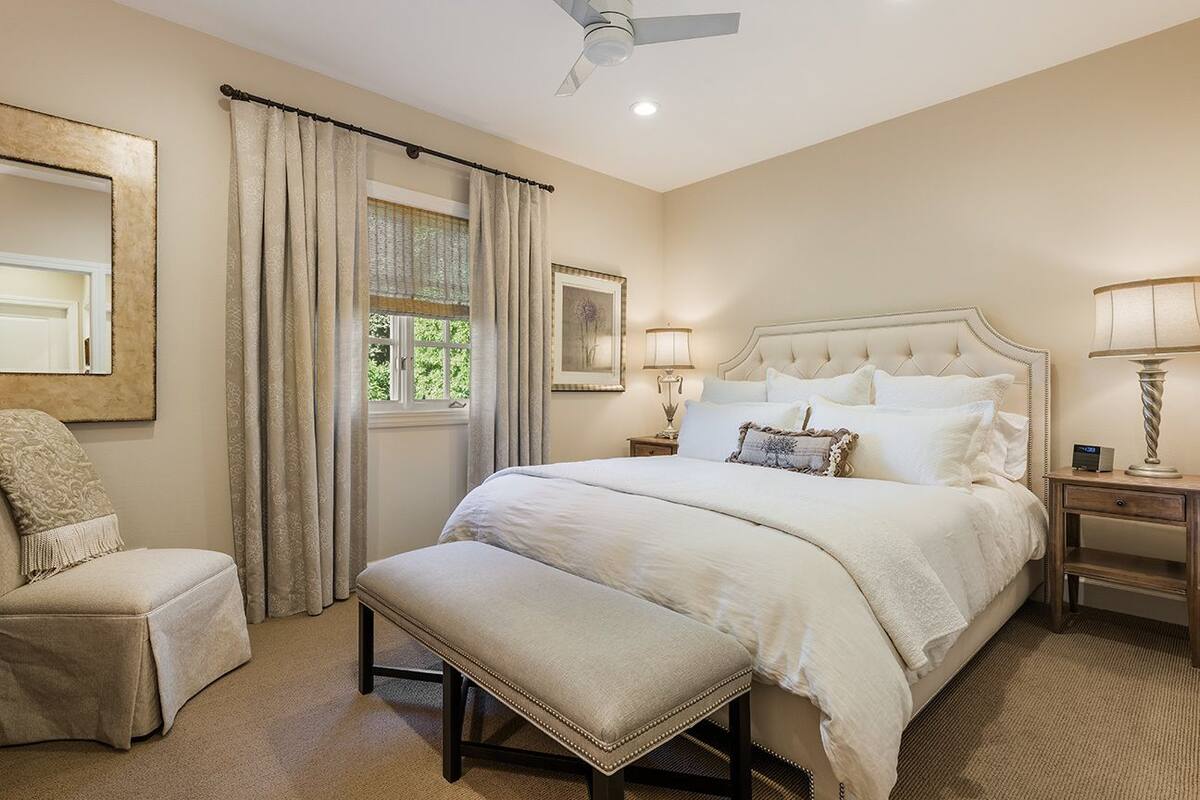 A bedroom with a large bed, a chair, a bench, a ceiling fan, and a window with curtains.