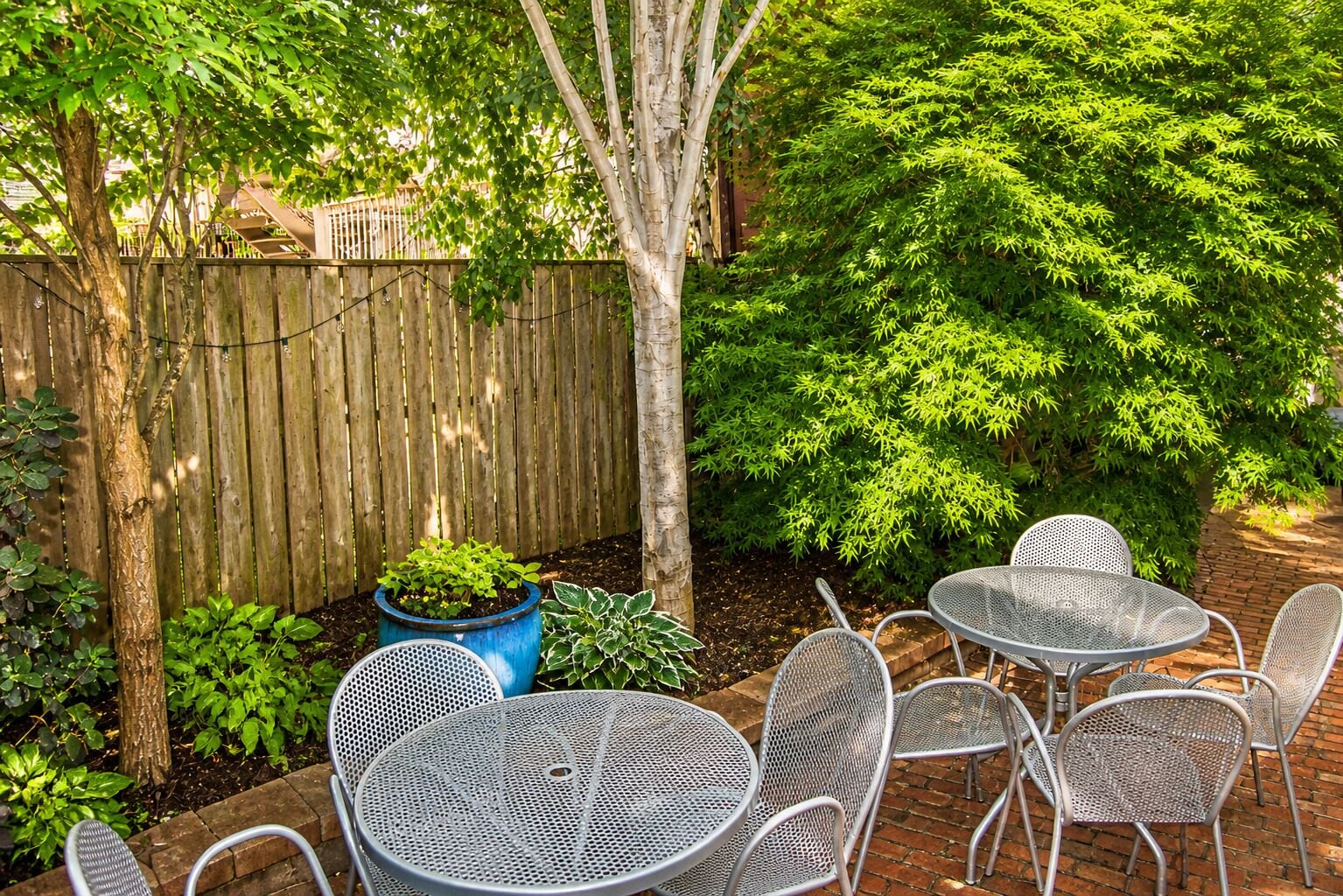 A patio with metal tables and chairs, surrounded by a wooden fence and lush greenery.
