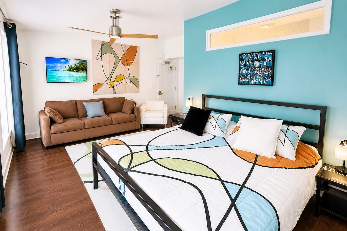 A bedroom with a bed, a sofa, a ceiling fan, a painting, and a framed photo on the wall.