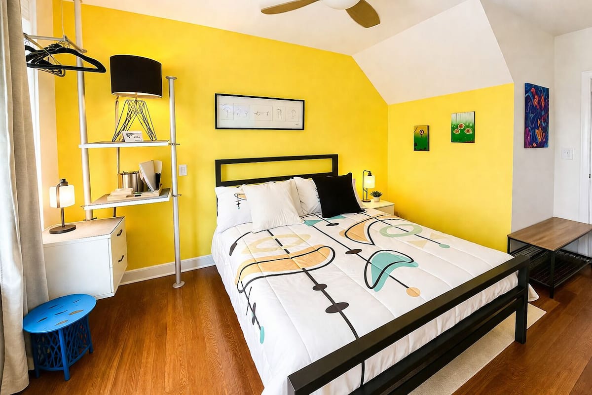 A bedroom with a bed, bedside tables, a desk, a chair, a hanging lamp, and artwork on the walls.