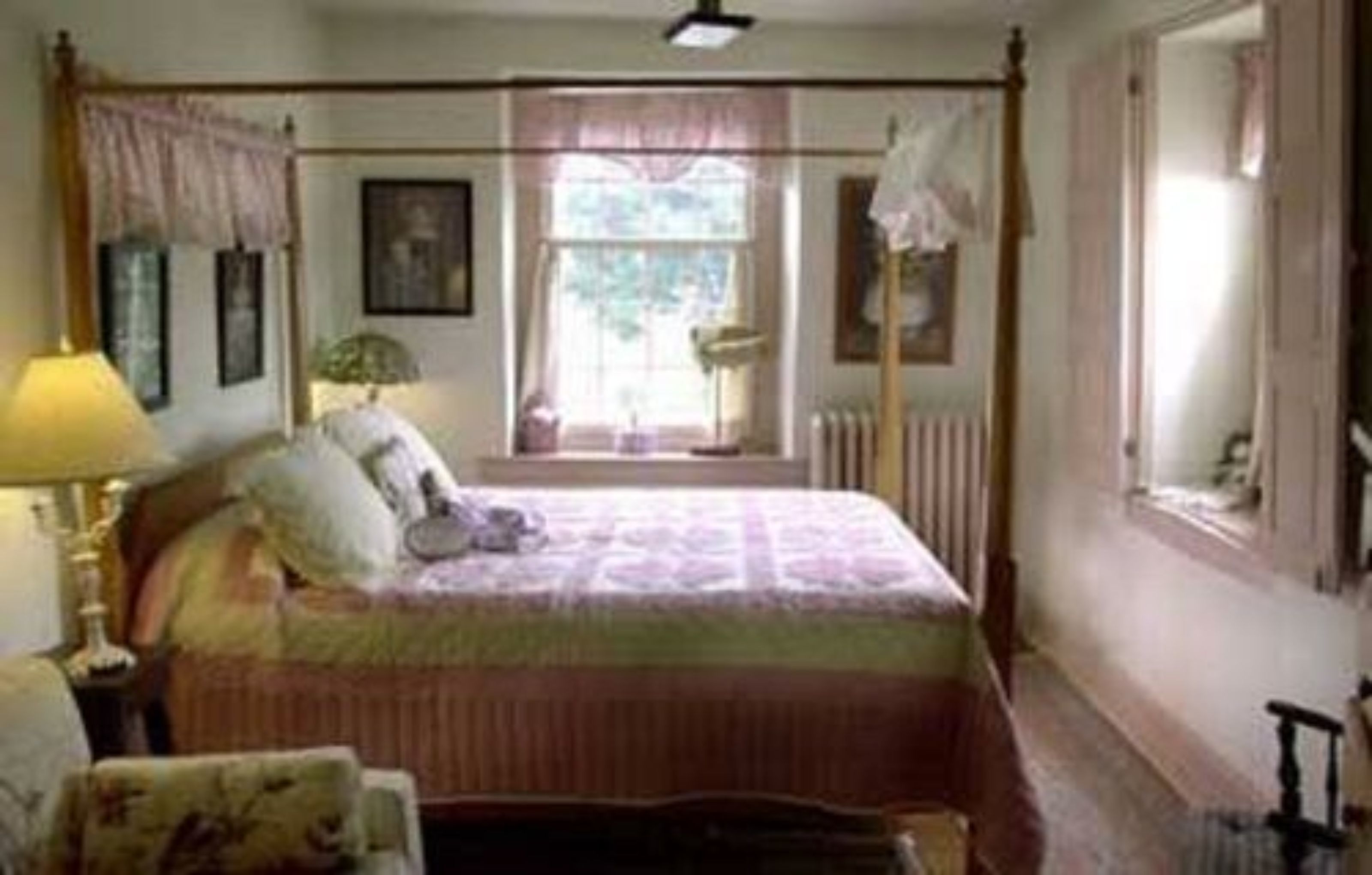 Ein traditionelles Schlafzimmer mit einem Himmelbett, einem Fenster mit Vorhängen, einem Heizkörper und einem Nachttisch mit Lampe.