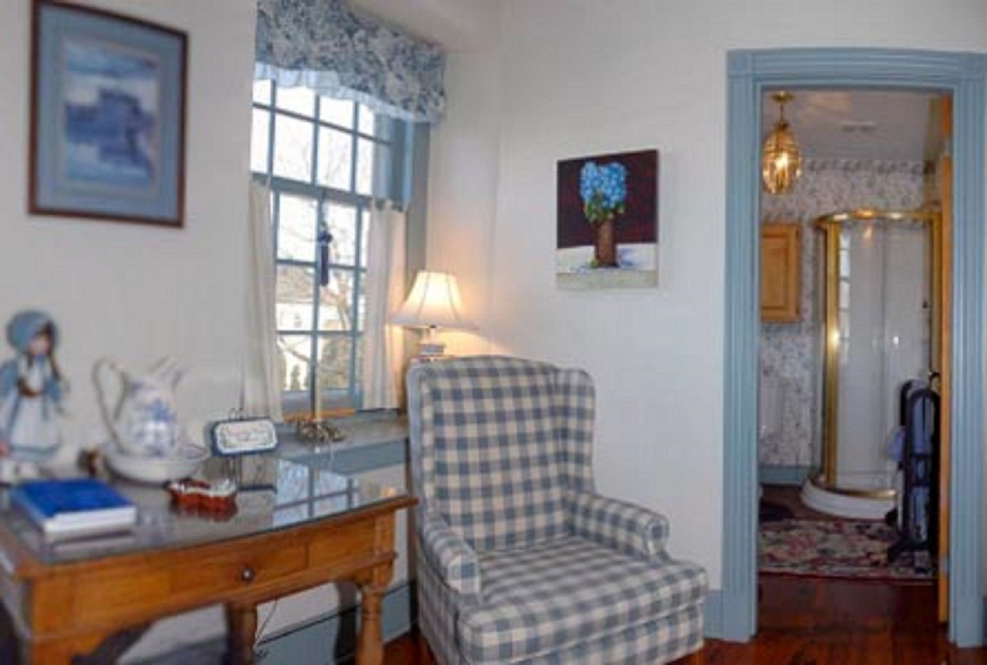Une pièce avec un fauteuil à motifs à carreaux, un bureau avec une lampe, un tableau encadré au mur et une fenêtre avec des rideaux.