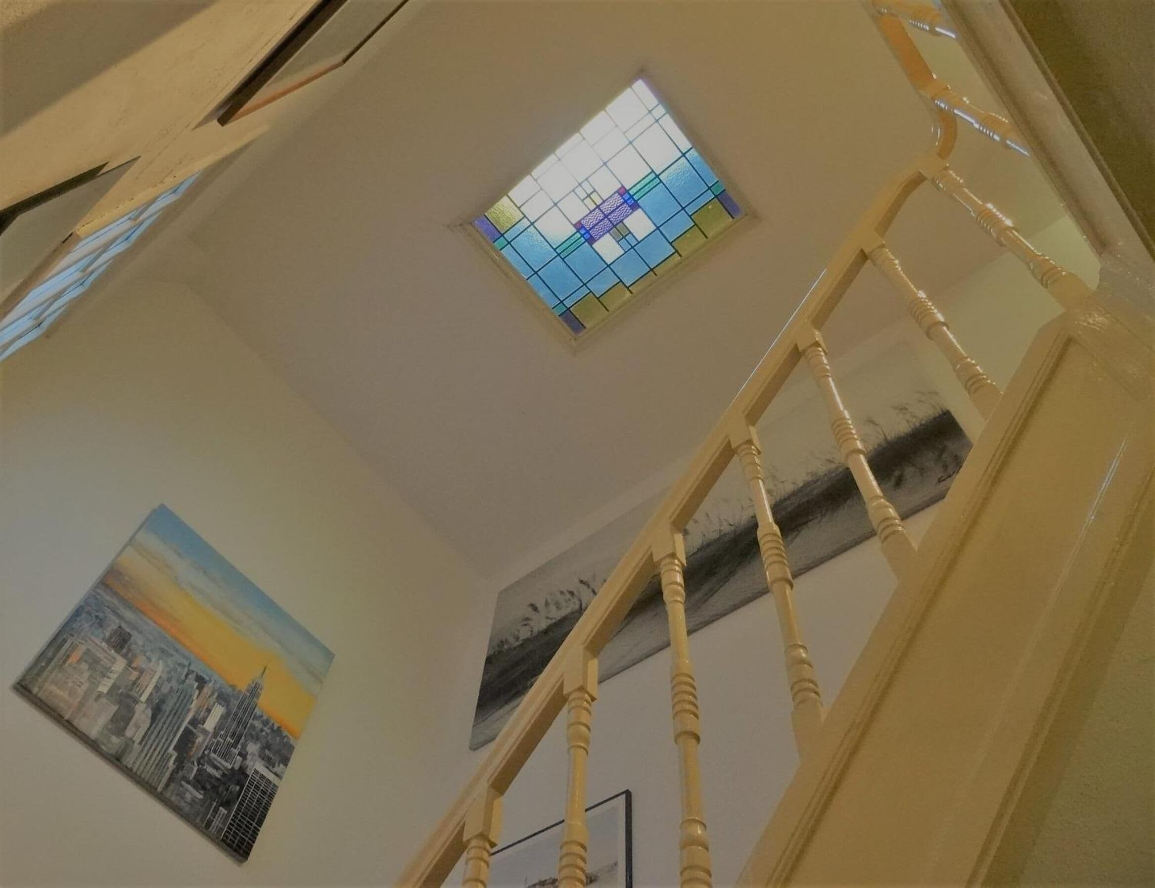Una escalera con barandilla de madera que lleva a una habitación con un cuadro de la ciudad y un vitral.