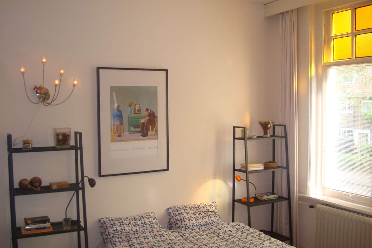 Un dormitorio con cama, dos estantes, un cuadro enmarco, un radiador y una ventana con cortinas.