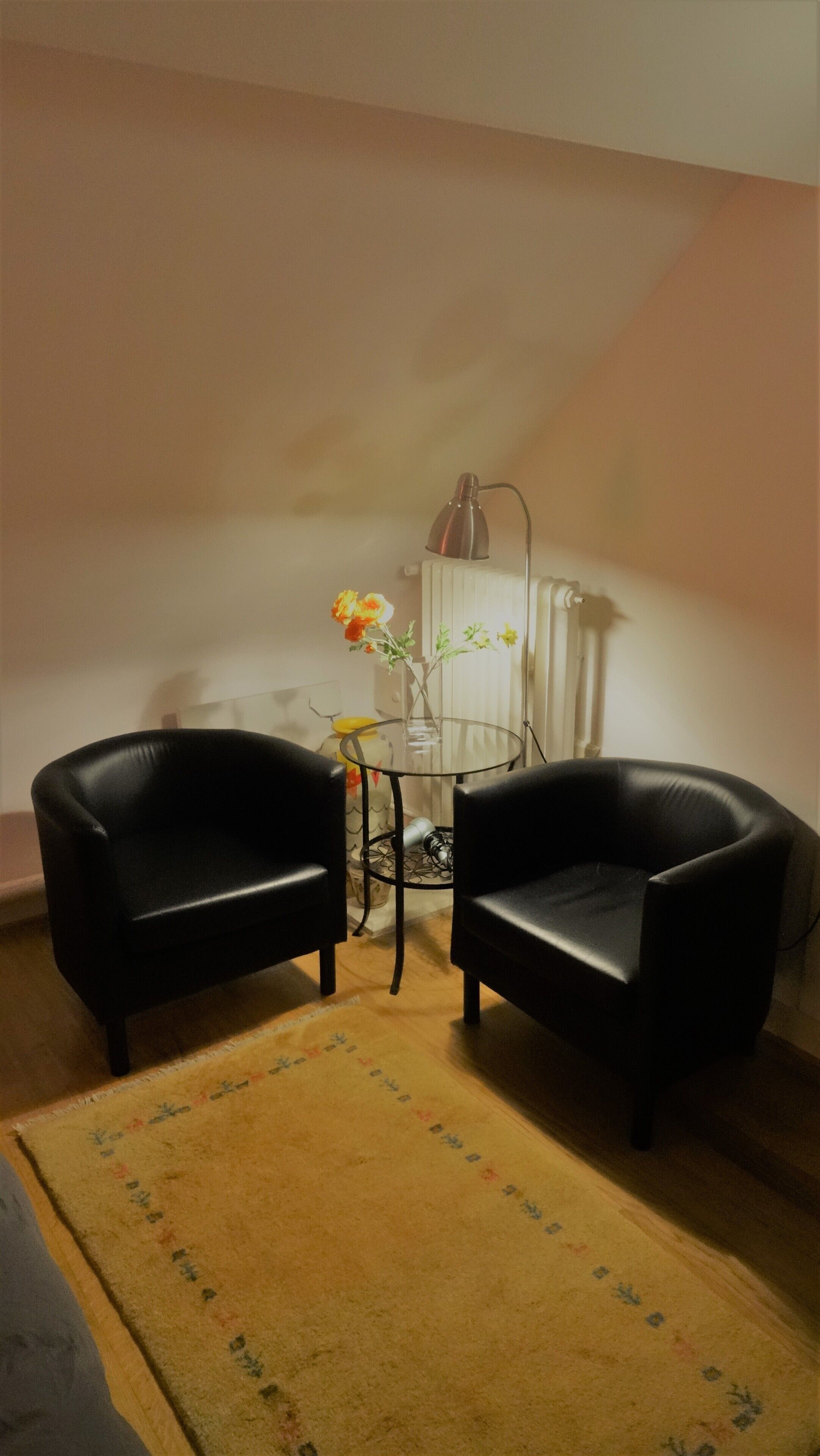 A room with two black leather chairs, a small table with a vase of flowers, a lamp, and a radiator.
