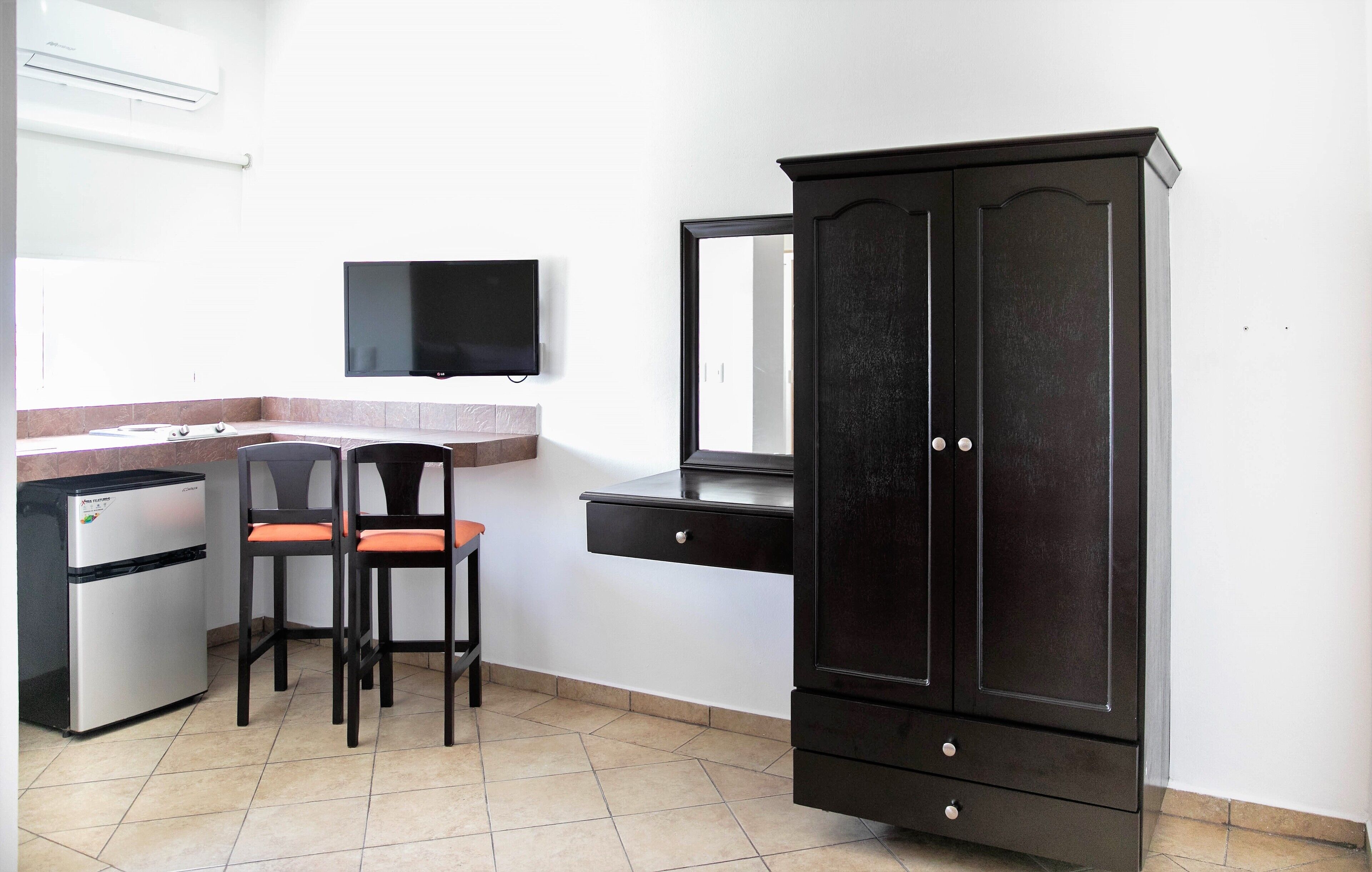 Una habitación compacta con kitchenette, televisor montado en la pared, mueble de madera oscura y una silla con cojines naranja.