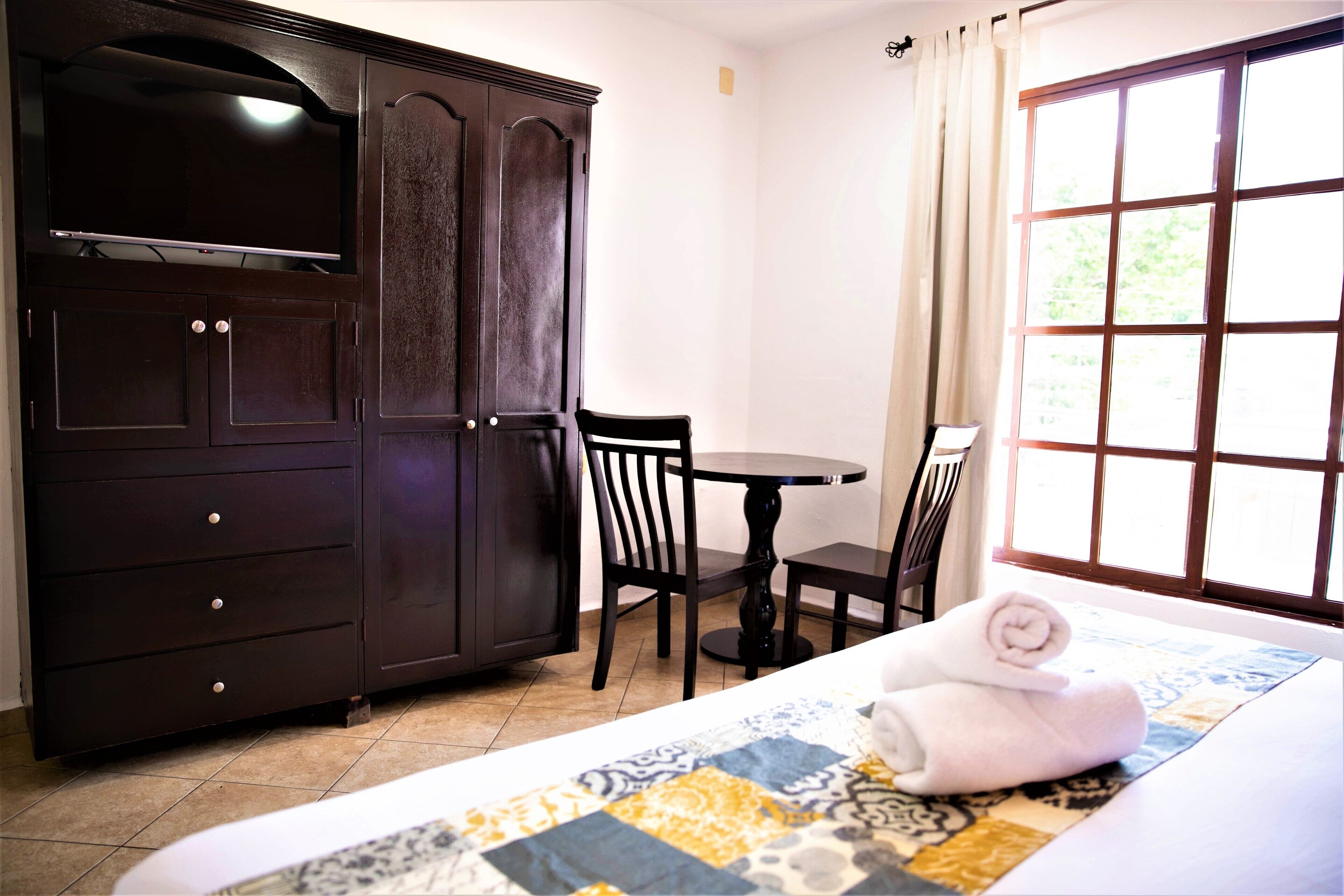 A hotel room with a bed, a dark wooden wardrobe, a small round table with two chairs, and a window with curtains.