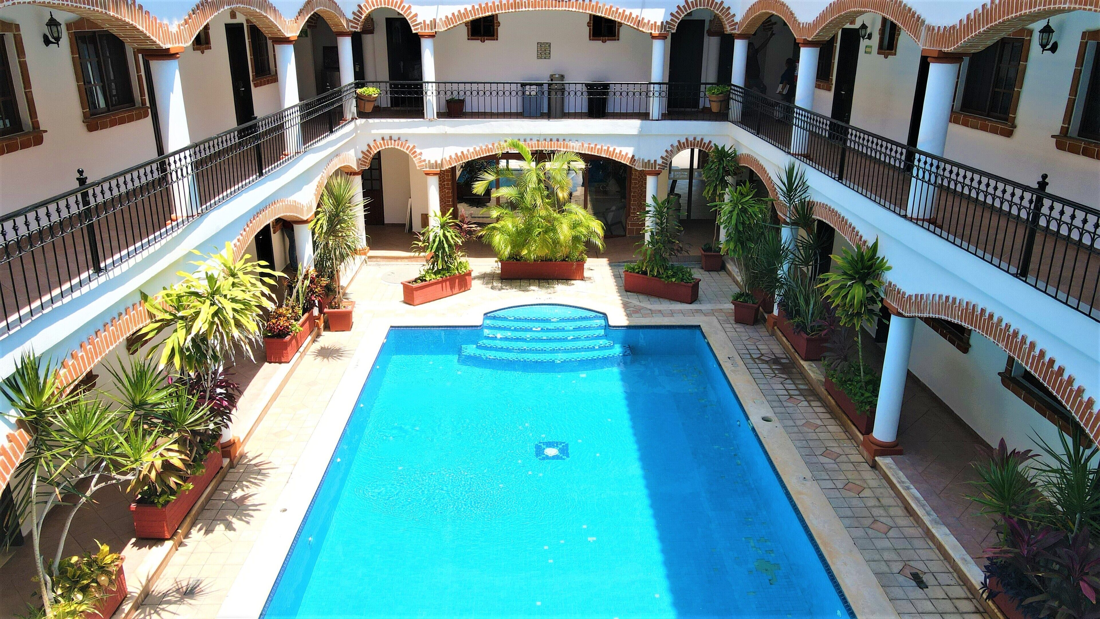 Um pátio com piscina, rodeado por plantas em vasos e um edifício de vários andares com varandas.