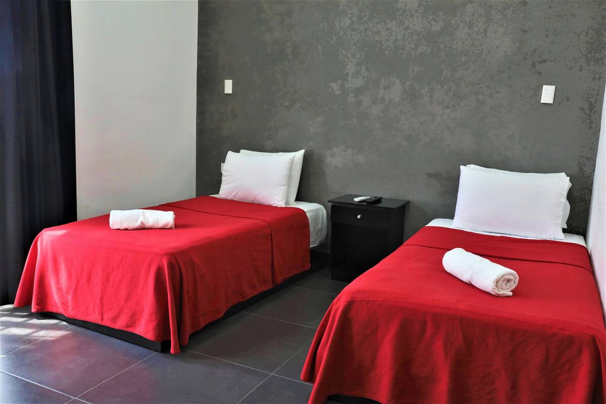 Dos camas individuales con ropa de cama roja, almohadas blancas y una mesita de noche negra con un control remoto en medio.