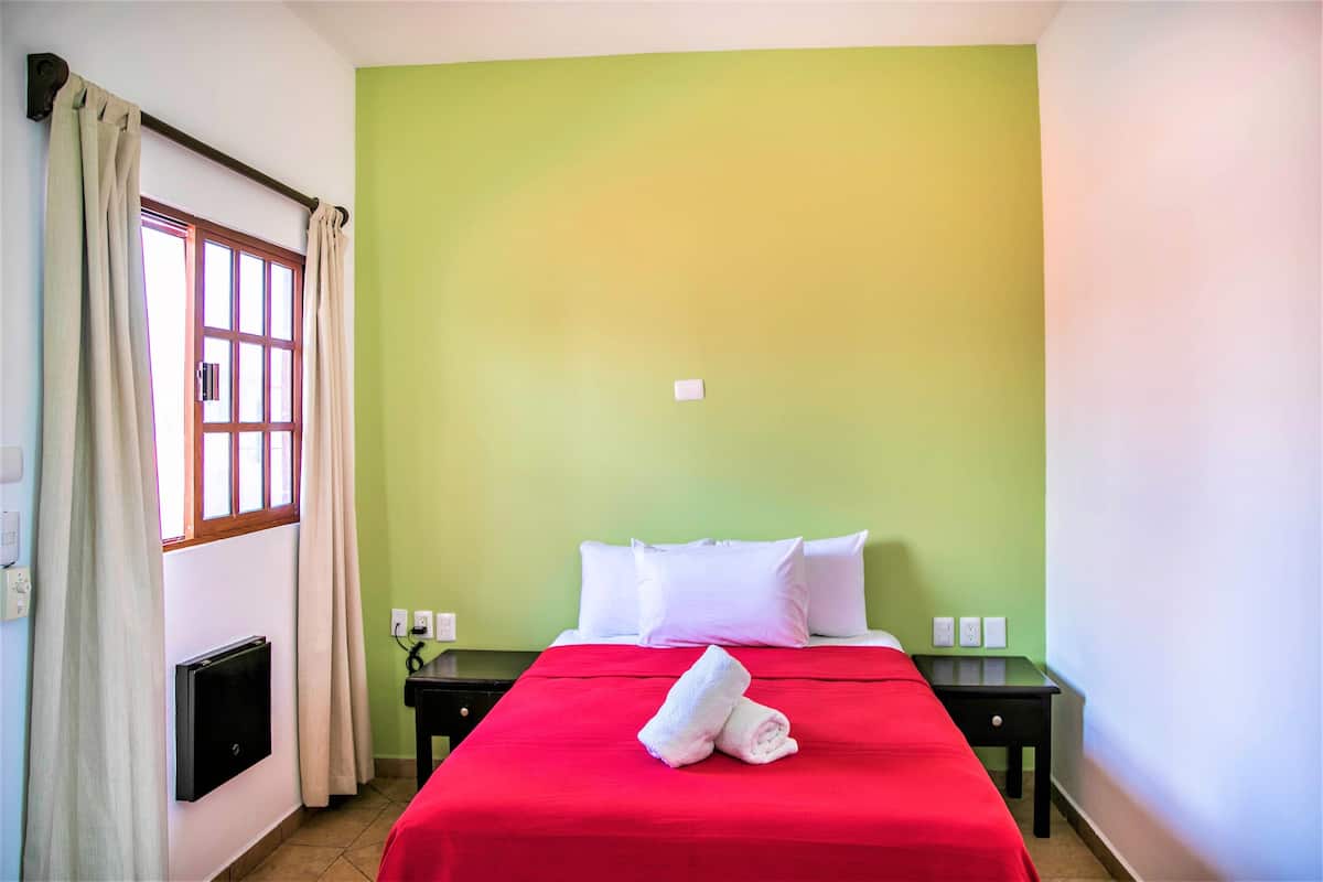 Una habitación de hotel con una cama, una mesita de noche, una ventana con cortinas y un termostato en la pared.