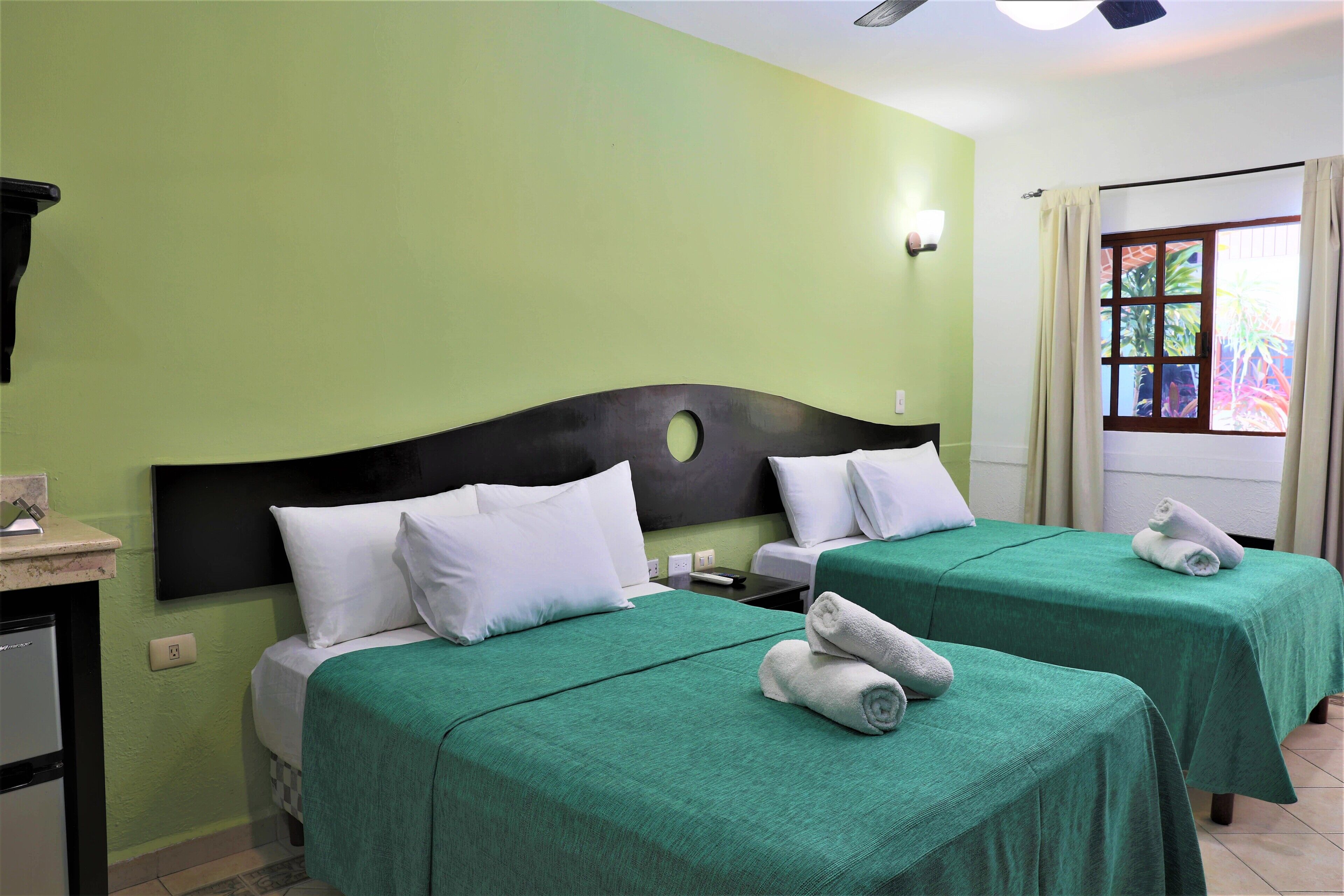 Une chambre d’hôtel avec deux lits, des couvre-lits verts, des oreillers blancs, un petit bureau avec un réfrigérateur et une fenêtre avec des rideaux.