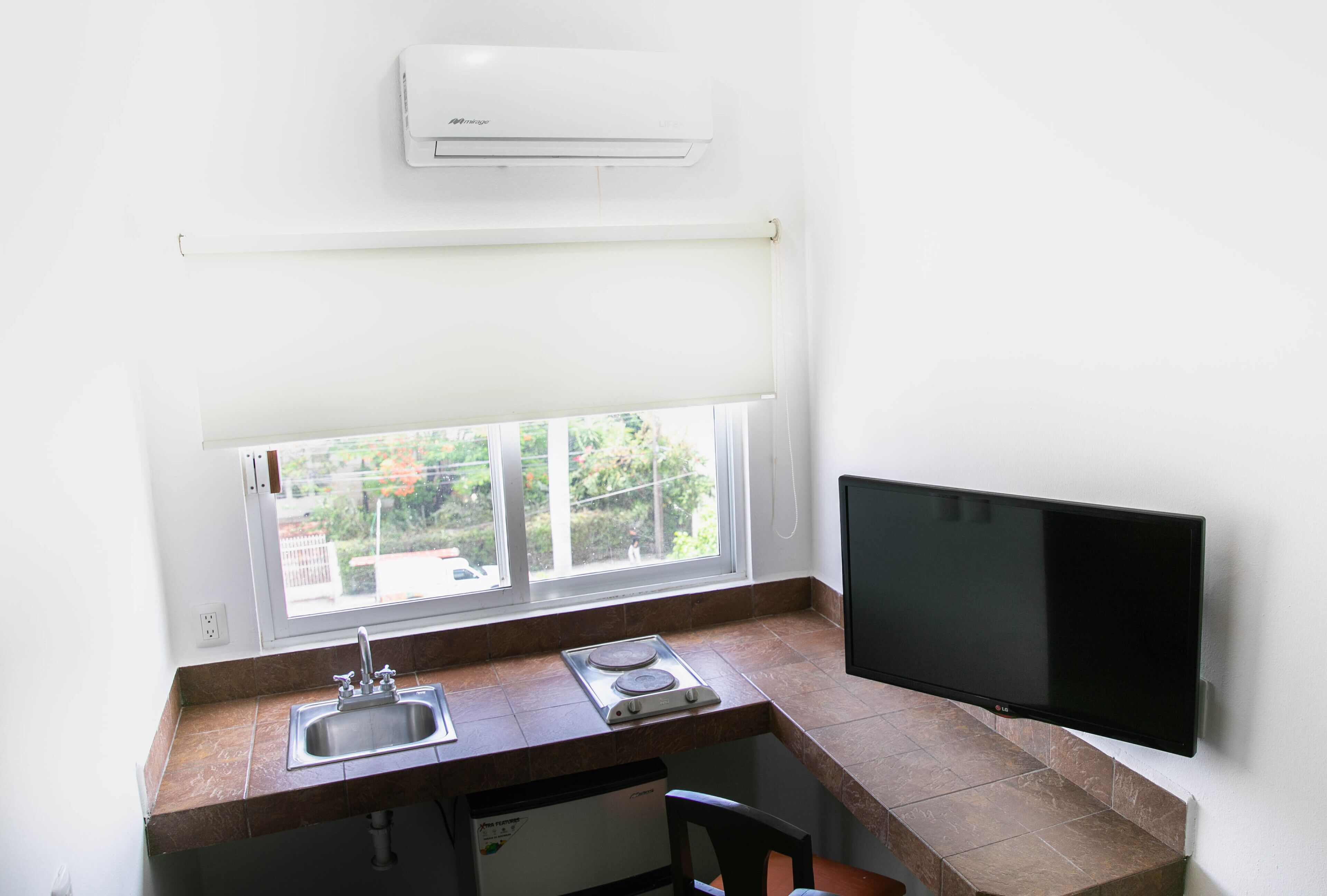 A small kitchen with a window, a sink, a stove, and a flat-screen TV mounted on the wall.