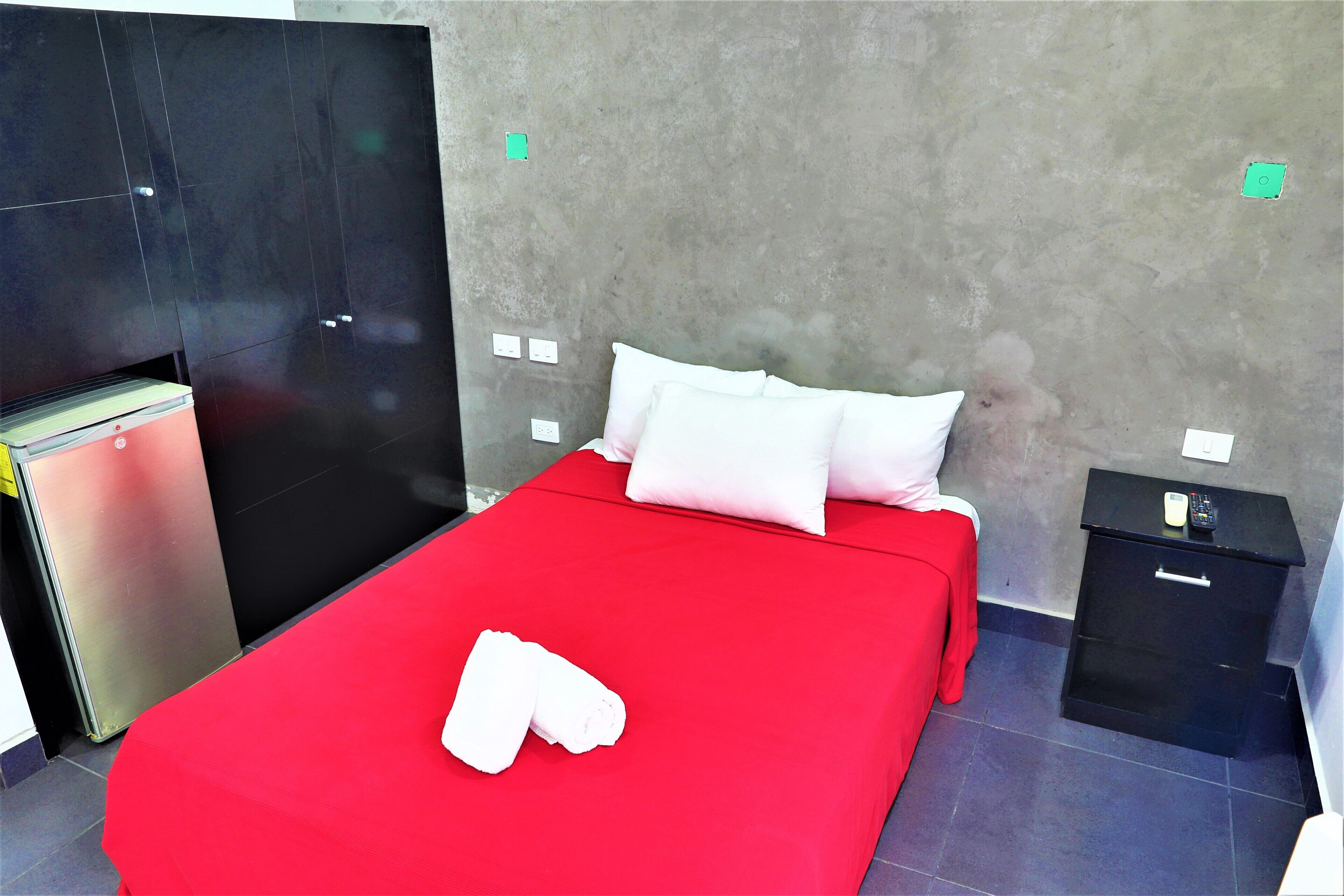 Quarto de hotel com cama vermelha, almofadas brancas, uma pequena mesa de cabeceira com um controle remoto e um criado-mudo preto.