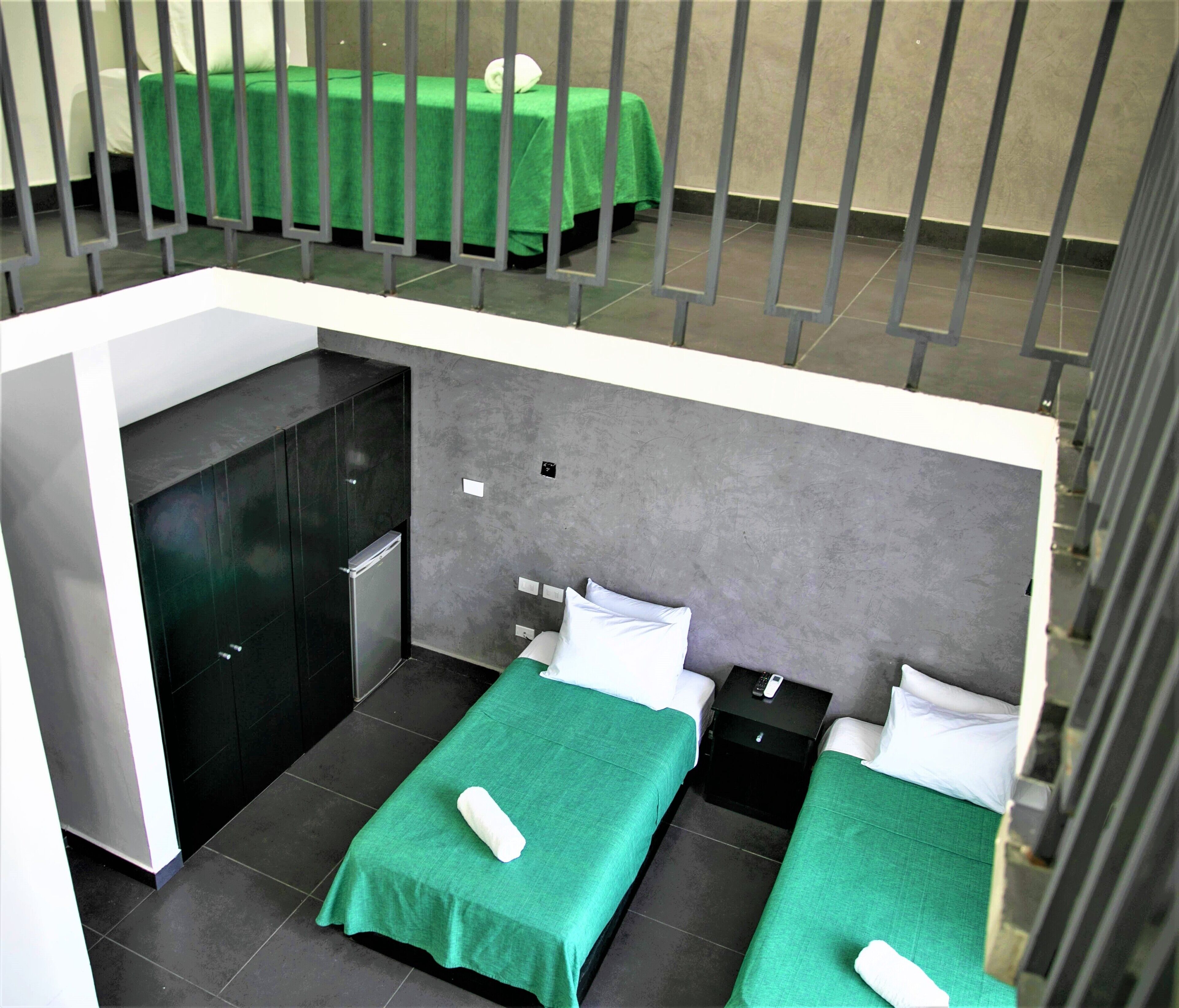 A hotel room with two beds, each with a green bedspread and white pillows. There is a small bedside table with a remote control on it. The room has a grey wall and a black cabinet.