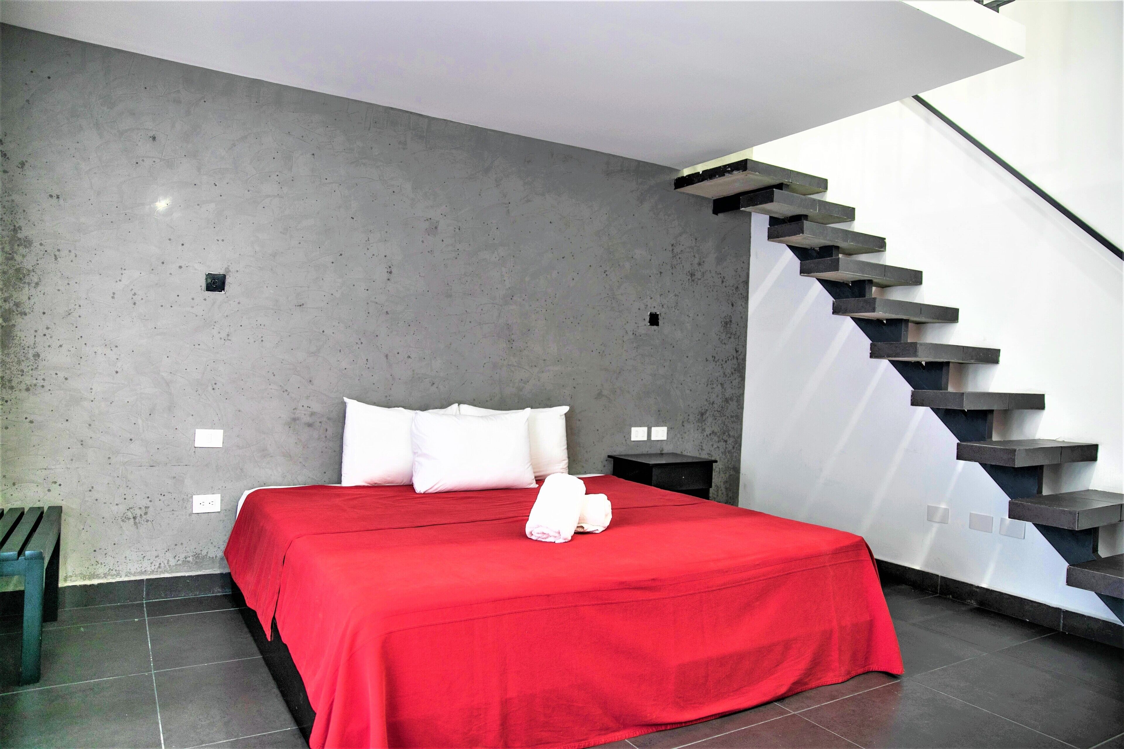 Quarto moderno com uma cama grande, um cobreleite vermelho, almofadas brancas, um banco de madeira e uma escada com degraus escuros.