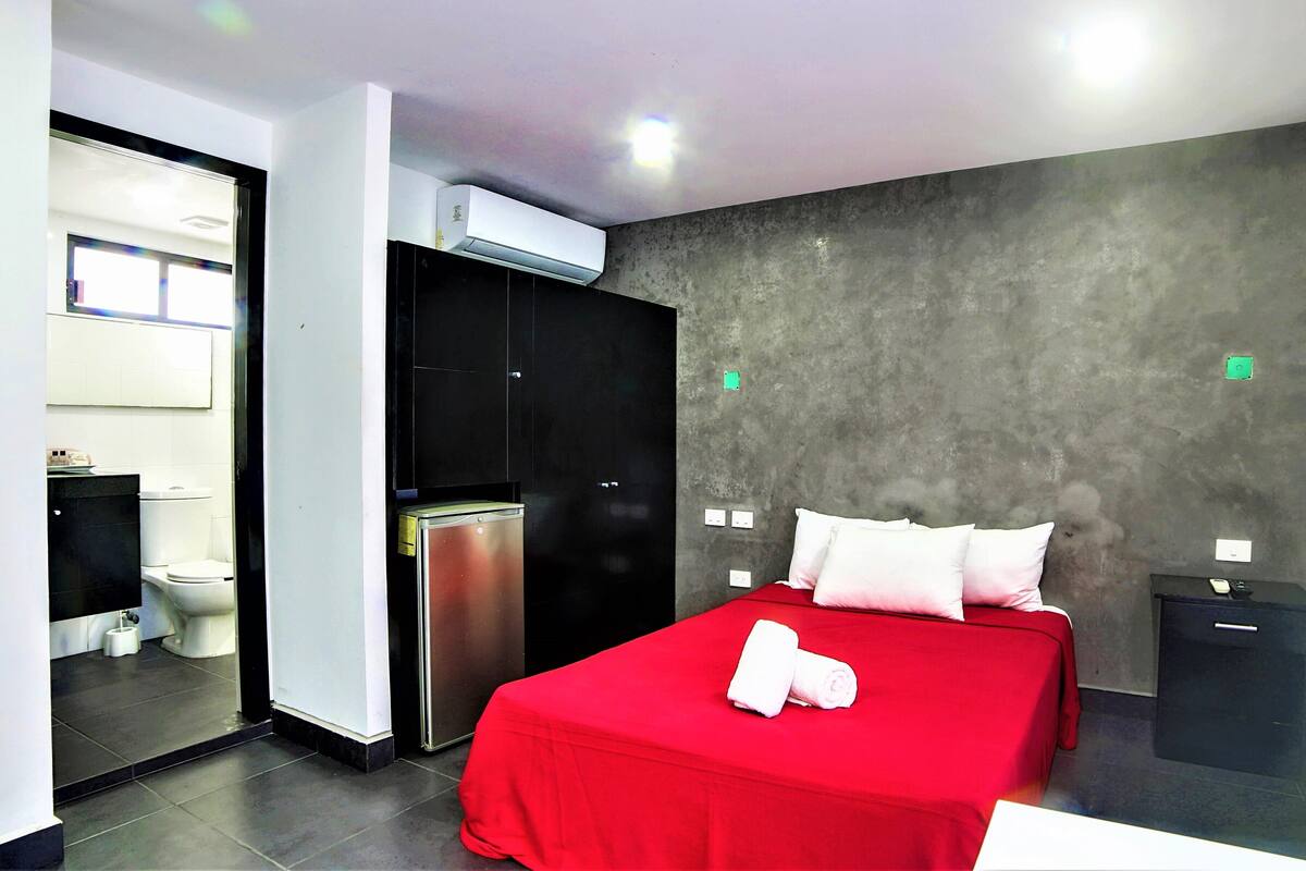 Habitación de hotel con cama roja, armario negro, inodoro blanco y pared gris.
