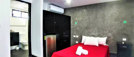 A hotel room with a red bed, a black wardrobe, a white toilet, and a grey wall.