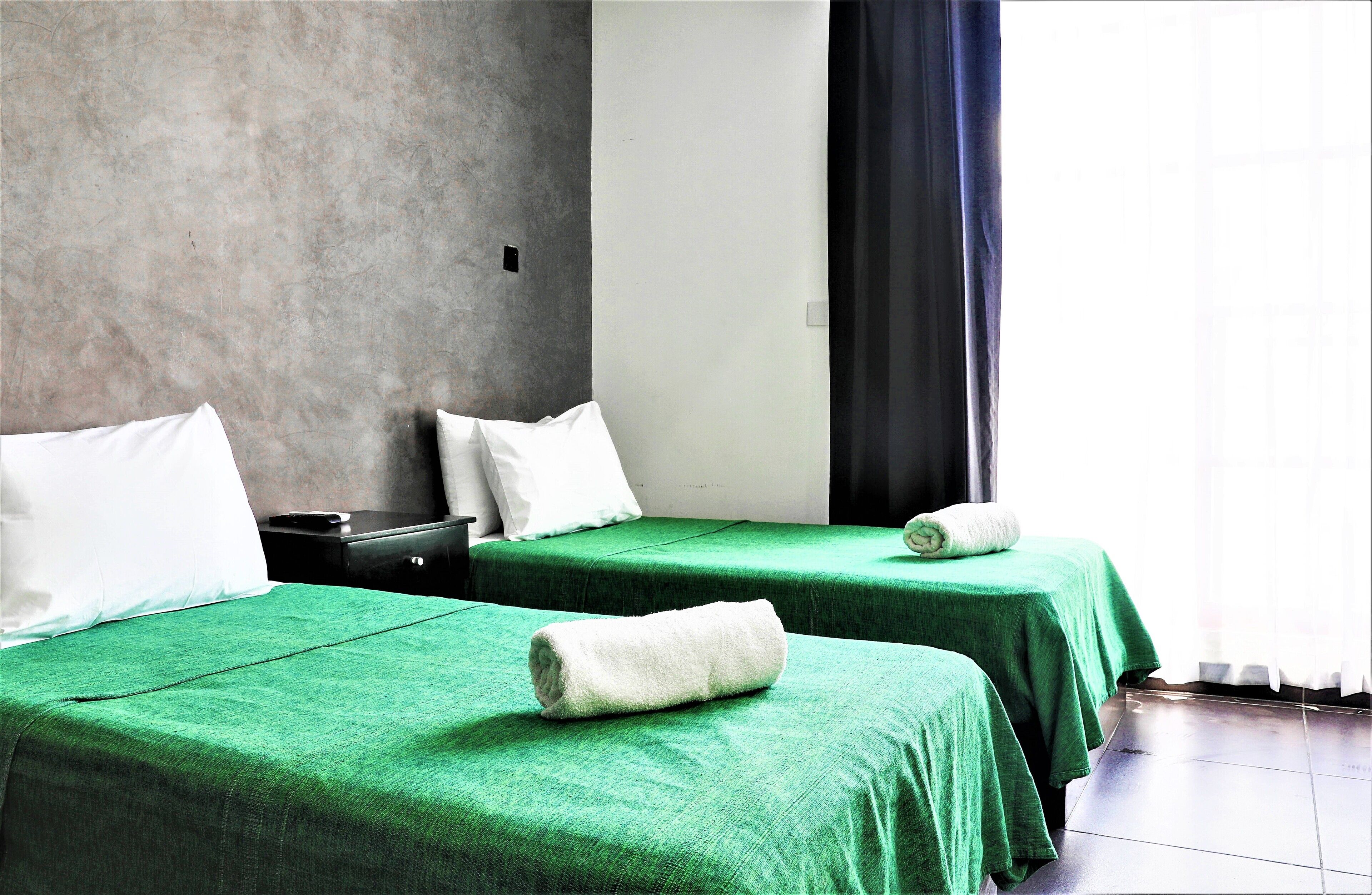 A hotel room with two beds, each with a green bedspread and white pillows. There is a small table with a black object on it, and a window with a dark curtain.