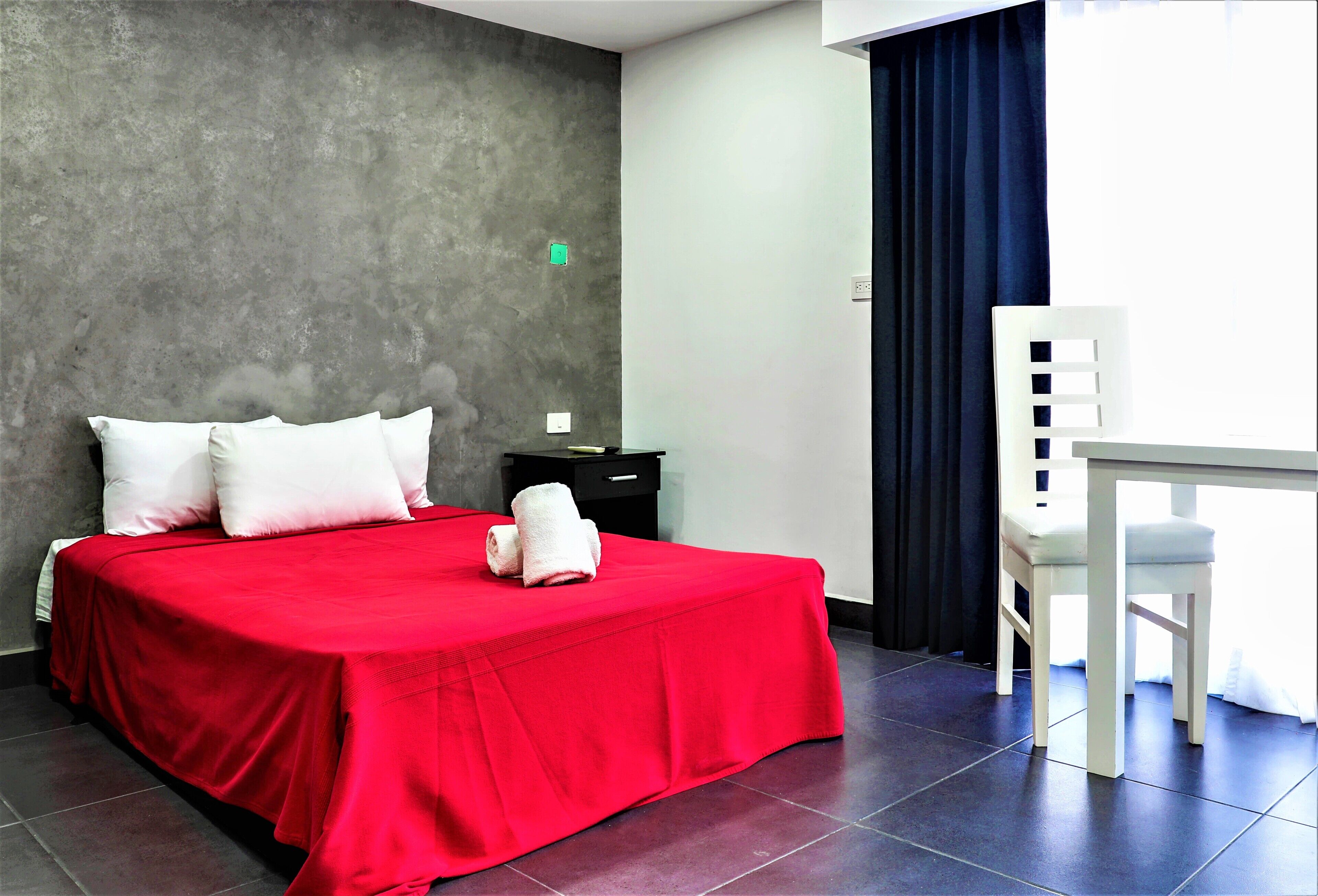 A hotel room with a red bedspread, white pillows, a white chair, a small table, and a dark grey textured wall.