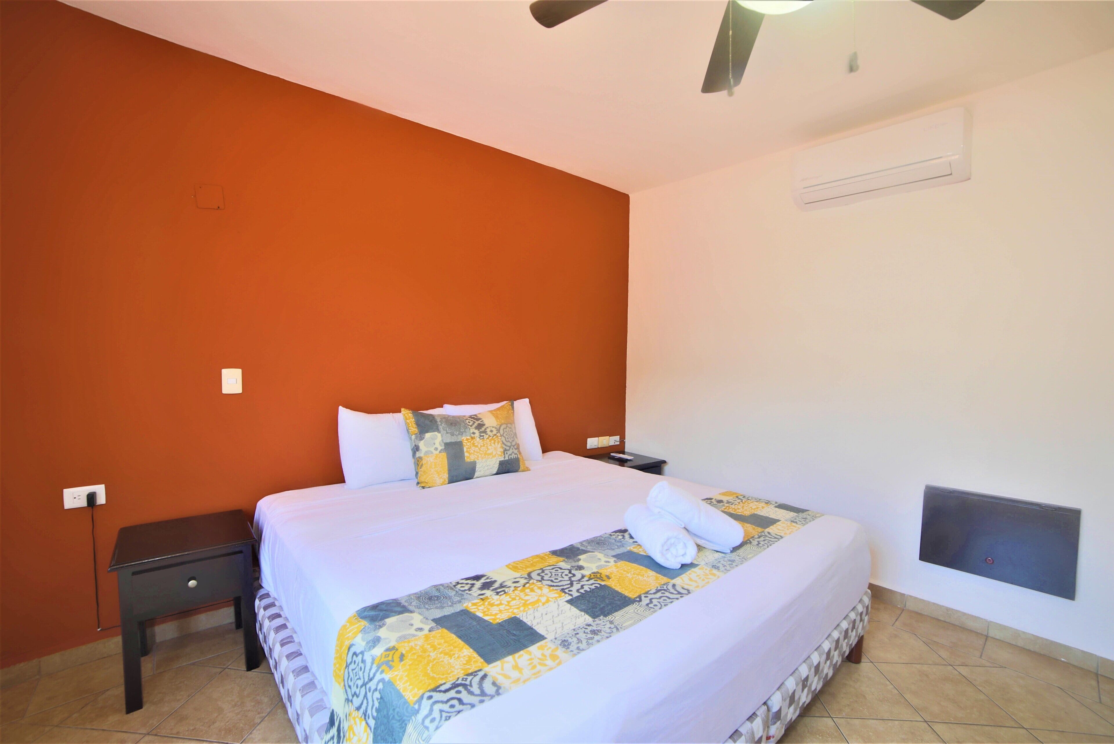 A neatly made bed with a patterned bedspread and two pillows, a bedside table with a lamp, and a wall-mounted air conditioner.