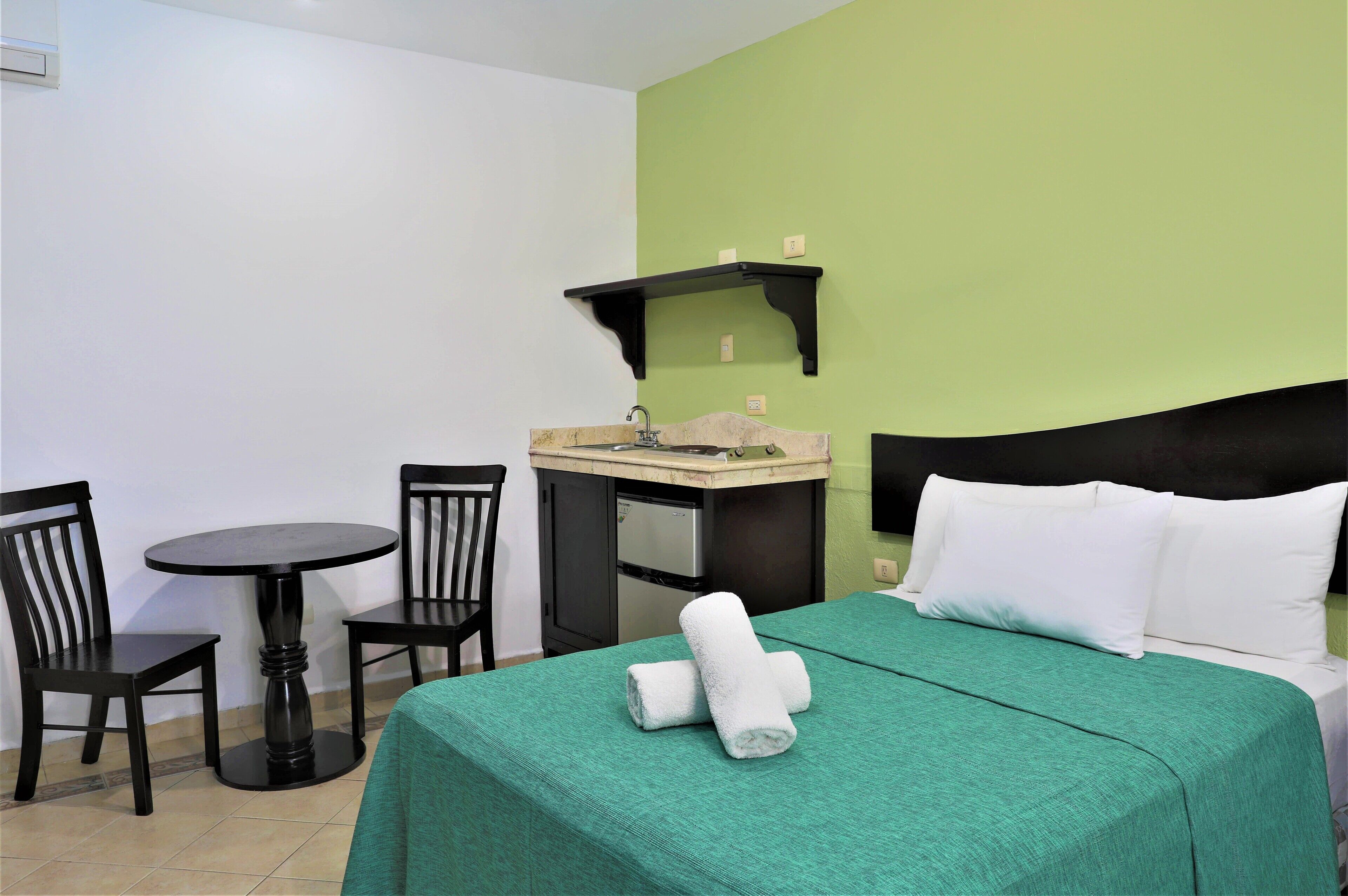 Habitación de hotel con cama, una mesita con sillas, kitchenette y pared verde.
