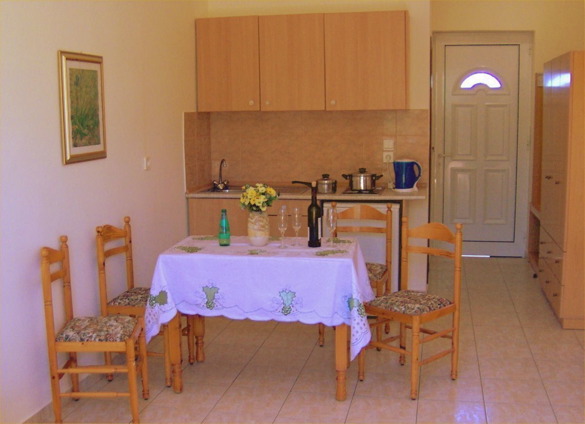 A dining area with a table set for four, wooden chairs, a white tablecloth, and a kitchen in the background with cabinets and a door.