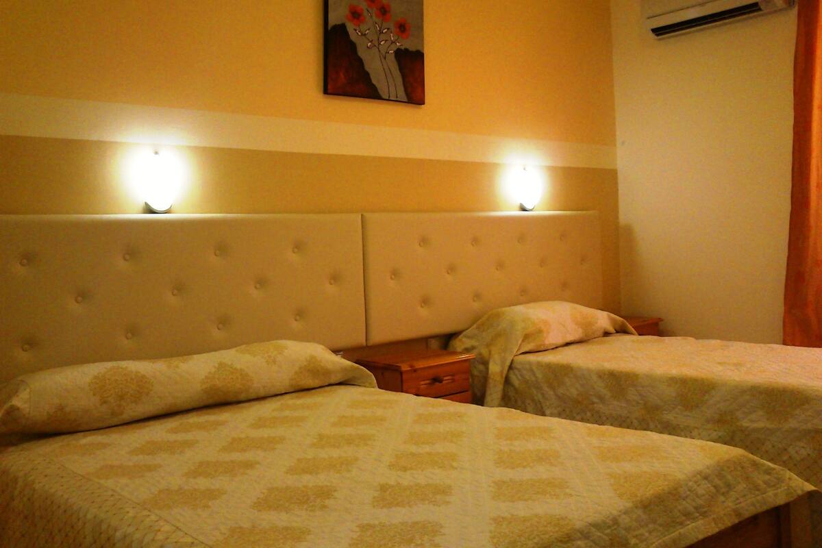 Een hotelkamer met twee bedden, een hoofdbord met verlichting, een airconditioner en een schilderij aan de muur.