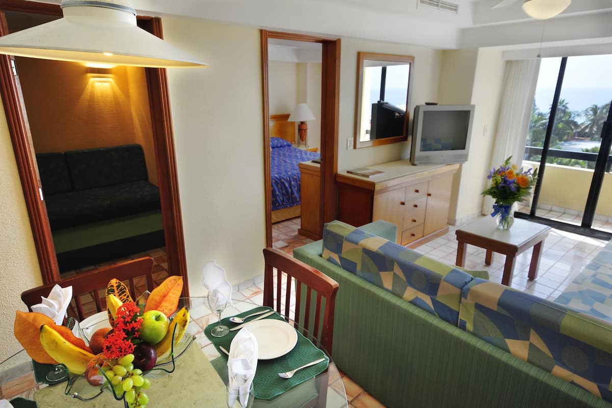 A hotel room with a dining area, a sofa, a television, and a balcony with a view.