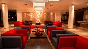 Eine moderne Lobby mit roten und grauen Sofas, die um einen zentralen Tisch angeordnet sind, großen Säulen und einer dekorativen Baumsulptur.