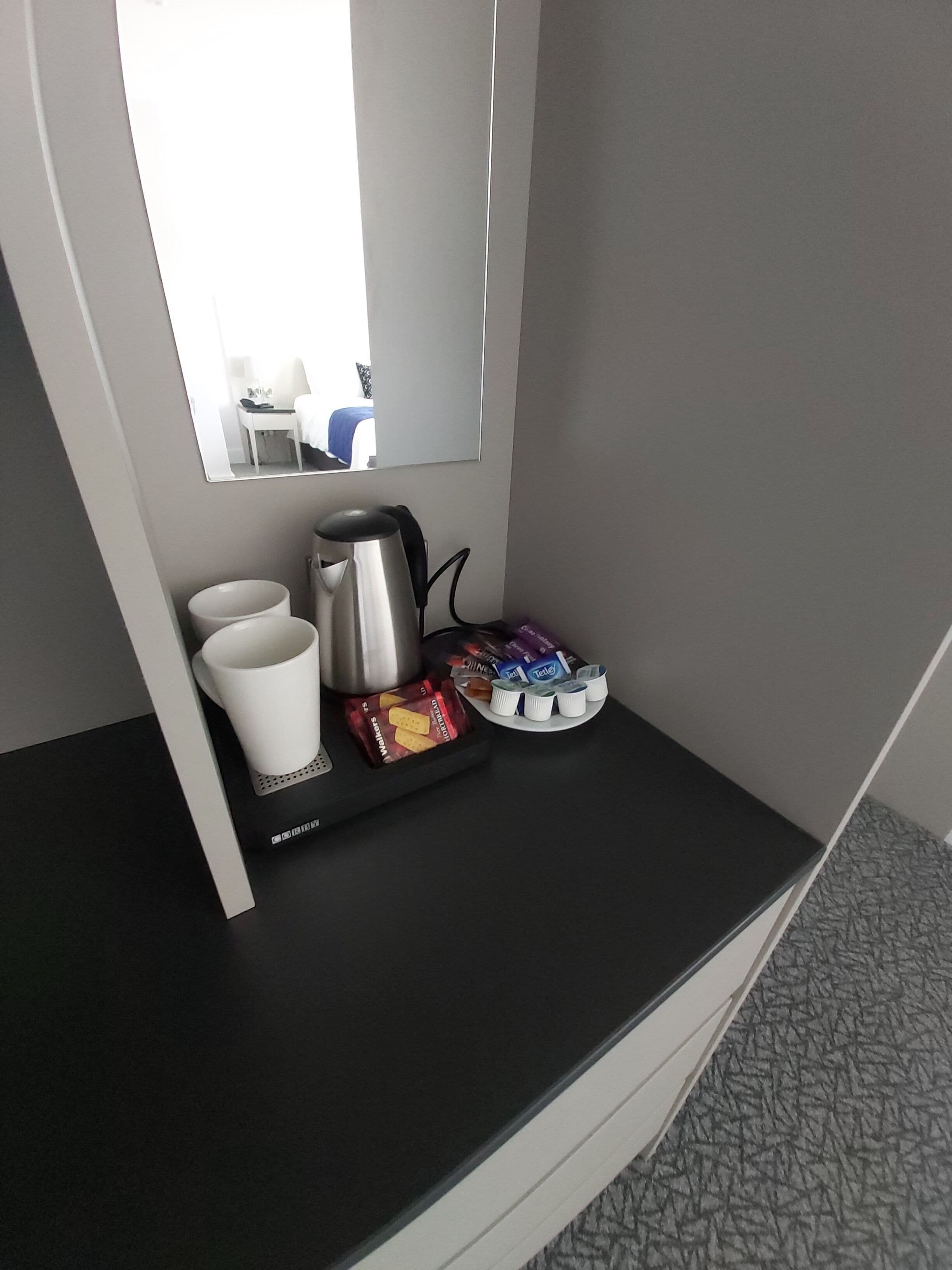 Une chambre d’hôtel avec une console noire, une tasse blanche, une bouilloire électrique argentée, un plateau avec des en-cas et un plateau de bouteilles d’eau.
