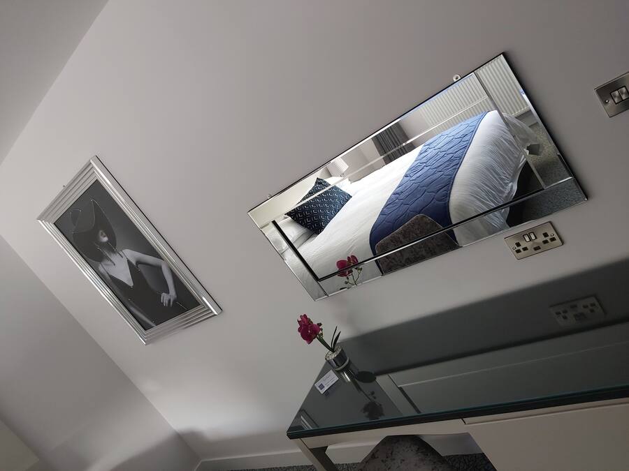 Un dormitorio con una cama, un espejo, una mesita con un florero y un enchufe de pared.