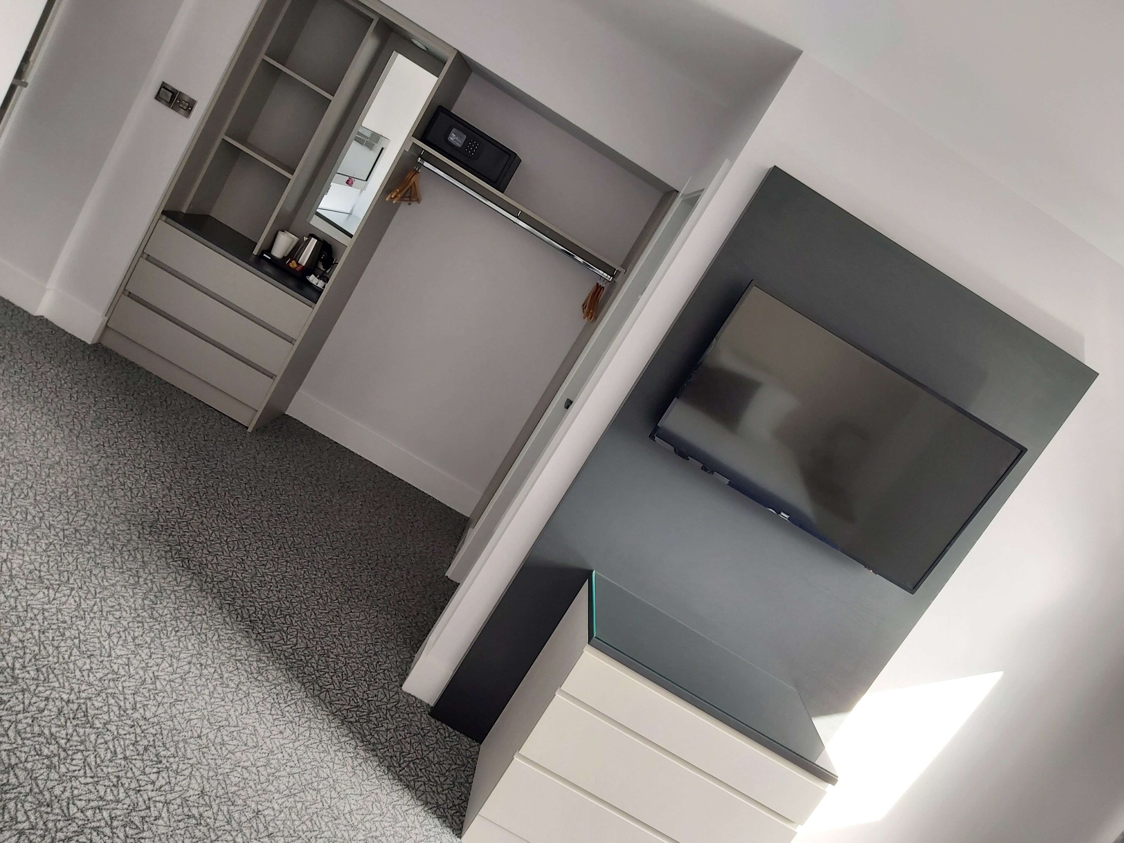 Una habitación de hotel moderna con vestidor, televisor de pantalla plana montado sobre un mueble blanco y alfombra estampada.