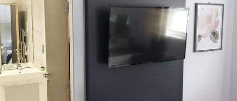 A bedroom with a bed, a television mounted on the wall, a bathroom with a mirror, and a door leading to another room.