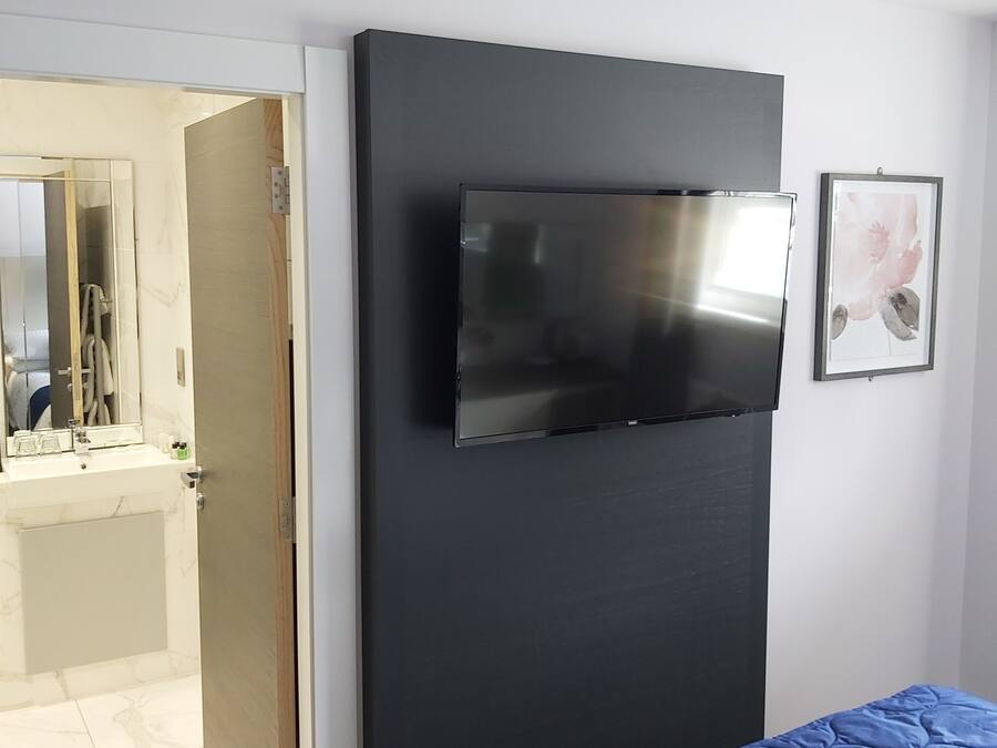 Quarto com cama, televisão na parede, banheiro com espelho e porta para outro cômodo.
