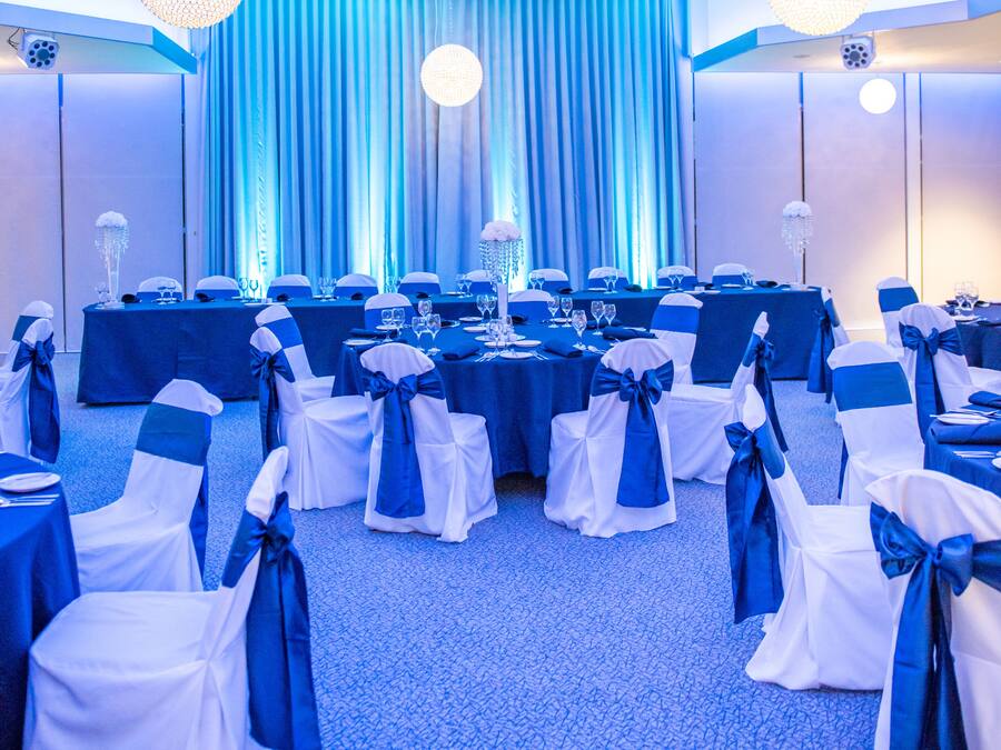Un salón de banquetes con mesas dispuestas para un evento formal, cortinas azules y candelabros.