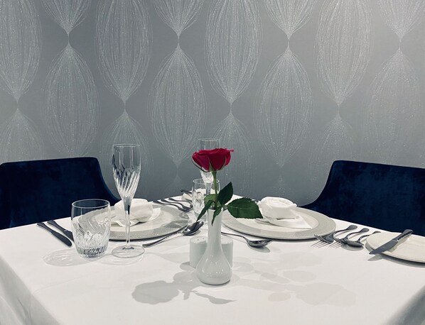 Una mesa para dos, con mantel blanco, un florero con una rosa roja y un fondo de papel tapiz estampado.